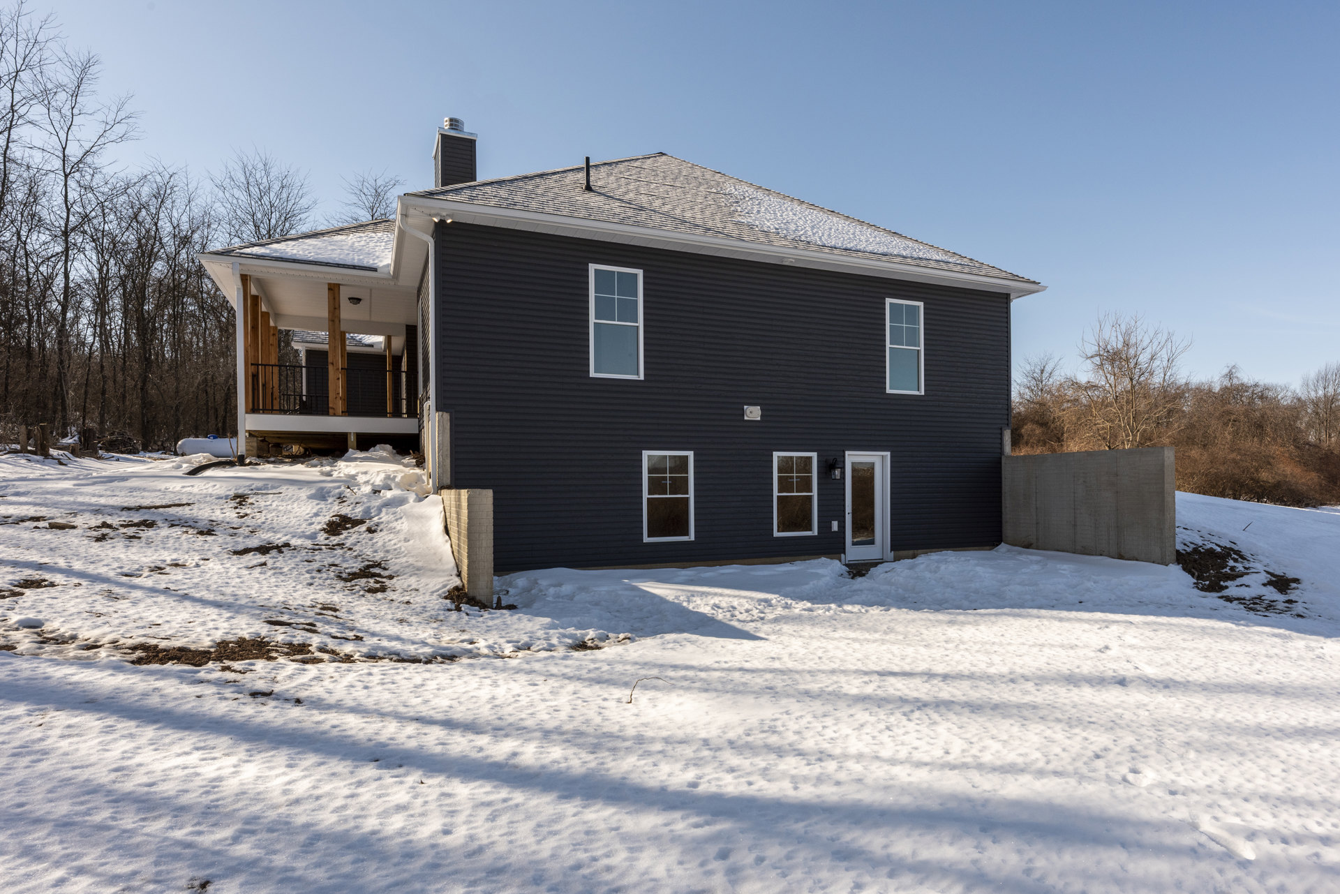Modern home exterior with white-framed windows, dark entry door, concrete walls dusted with snow, leafless trees in the yard, and snow-covered ground