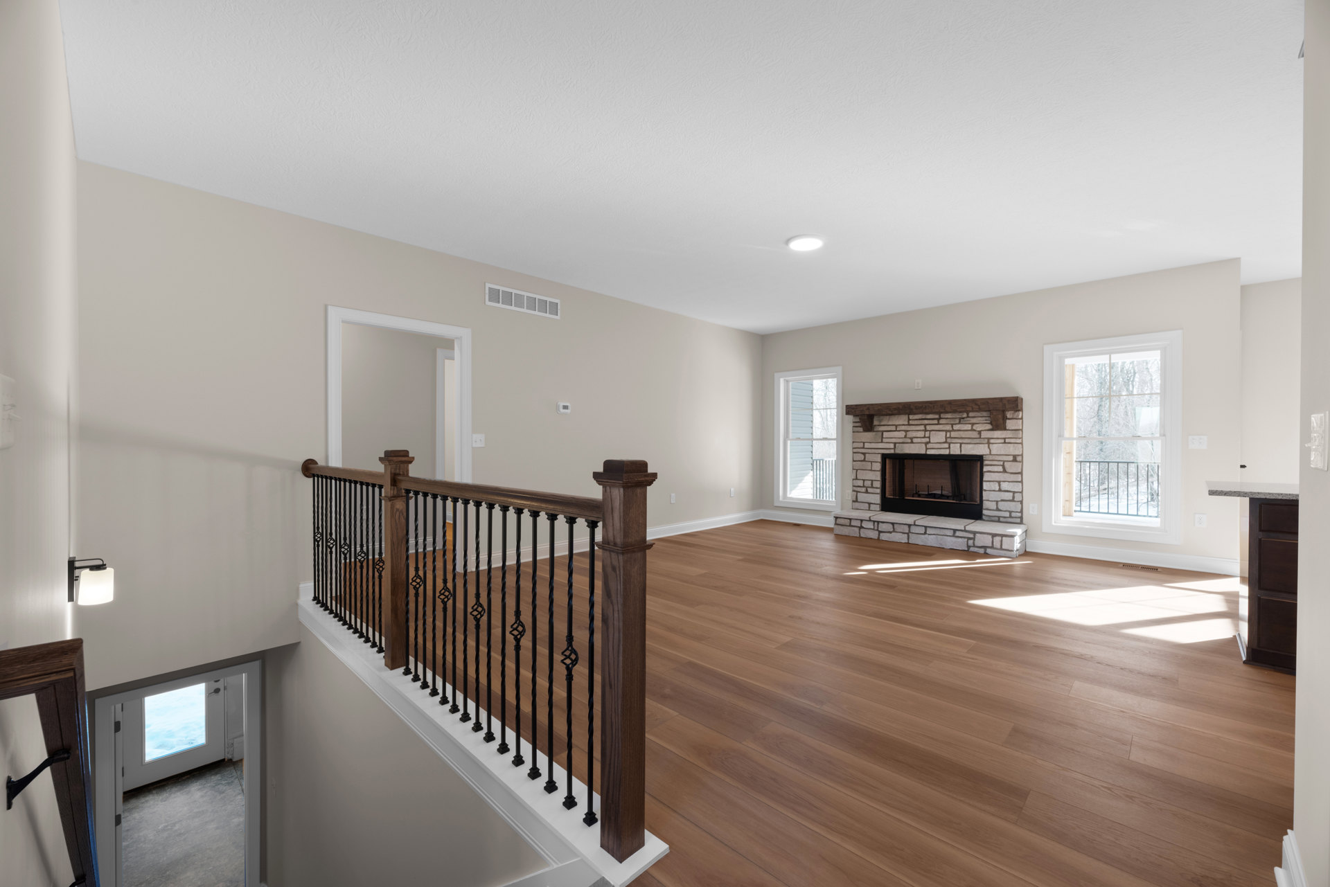 Living room with hardwood floors, brick fireplace featuring a wood beam mantel, large windows overlooking snowy trees, and a close-up of a modern railing.