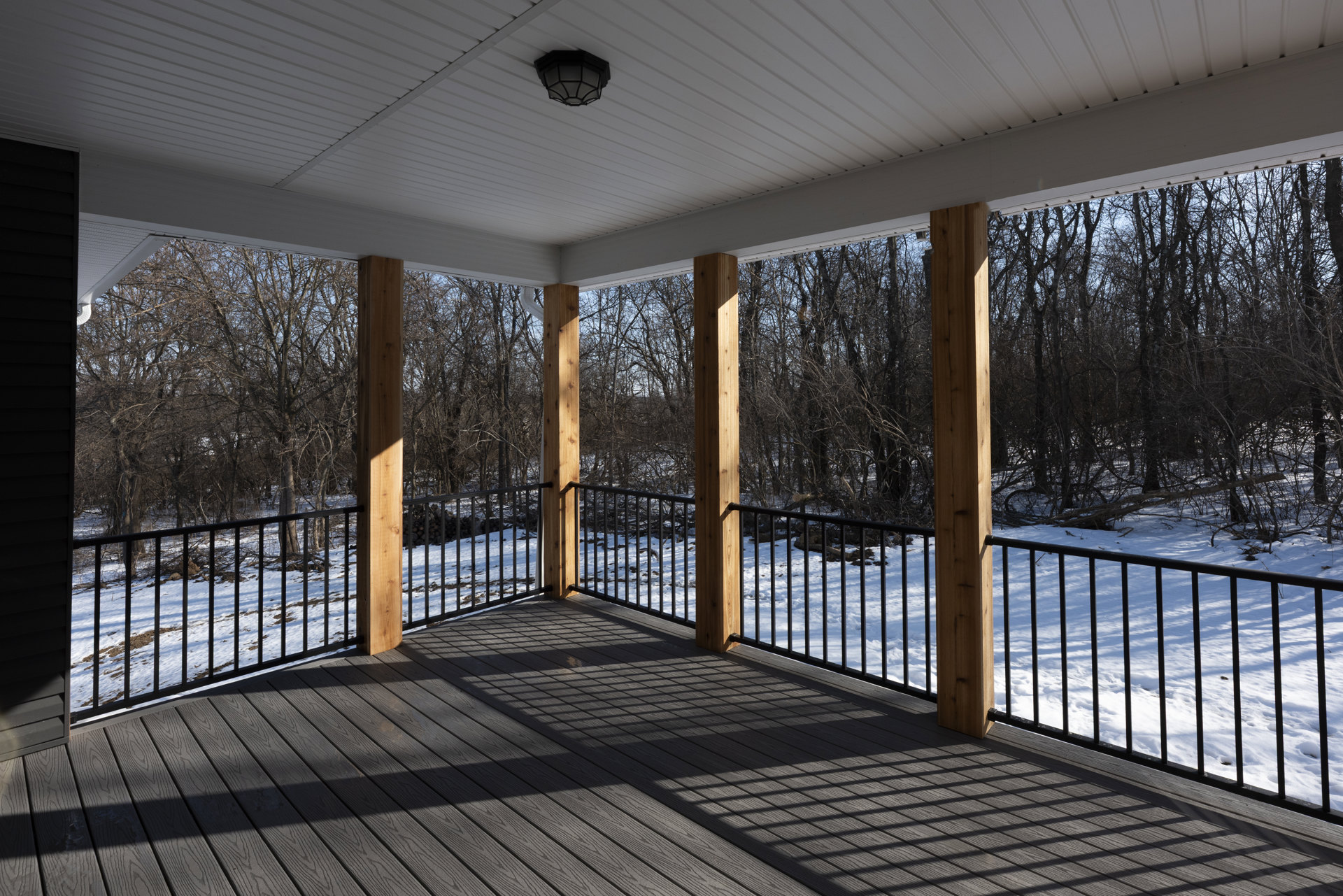 Wood deck with black metal railing, snow covering the ground, bare trees in background, covered porch ceiling with recessed light fixture visible