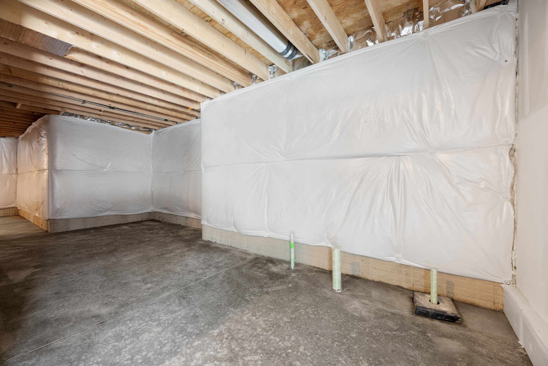 Concrete floor with white support poles, white plastic sheeting covering walls, exposed pipe in floor opening, black rectangular object with hole, grey tile flooring with visible