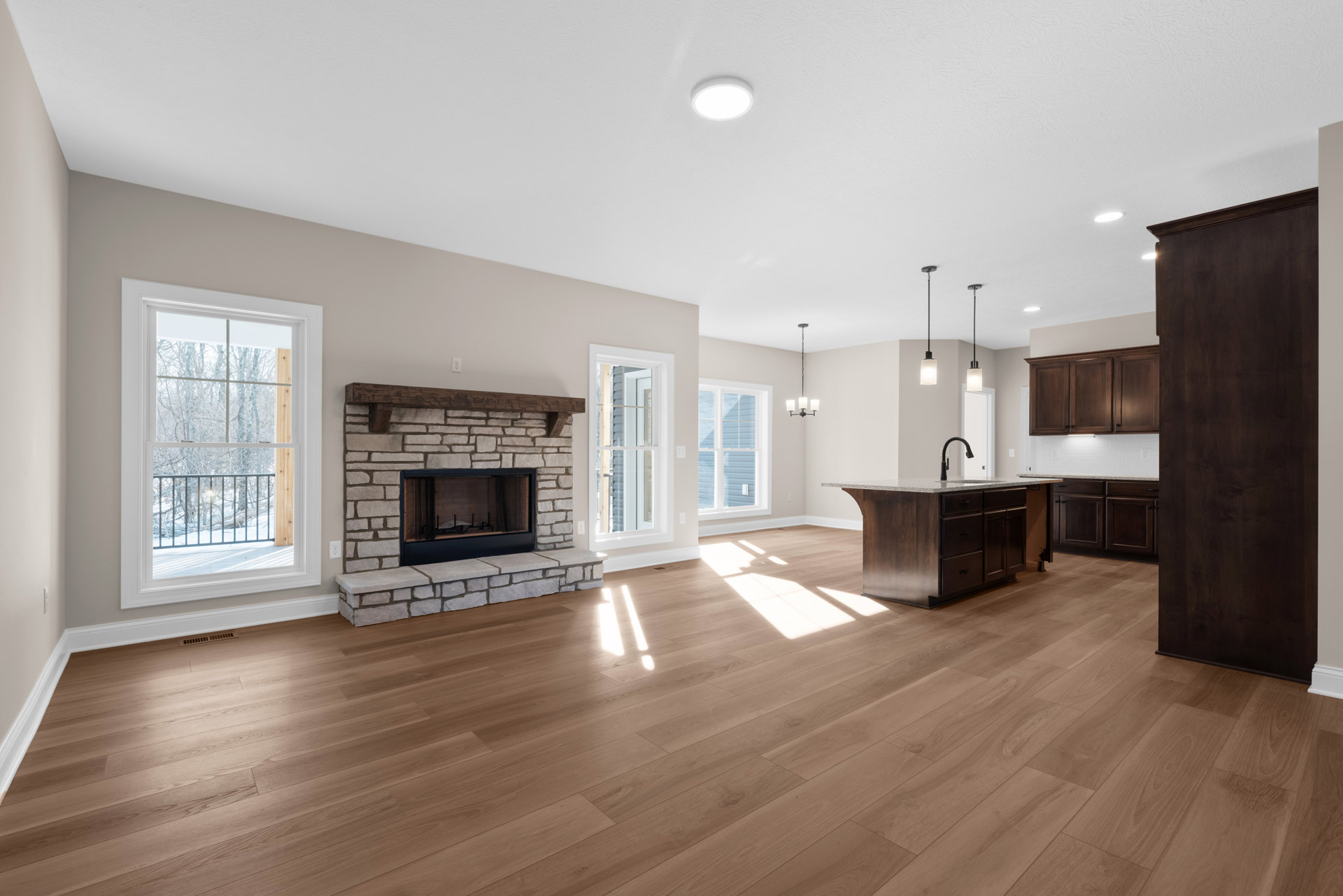 Spacious open floor plan featuring hardwood flooring, dark wood paneled wall, central fireplace with black mesh curtain, adjoining kitchen, large window with railing overlooking