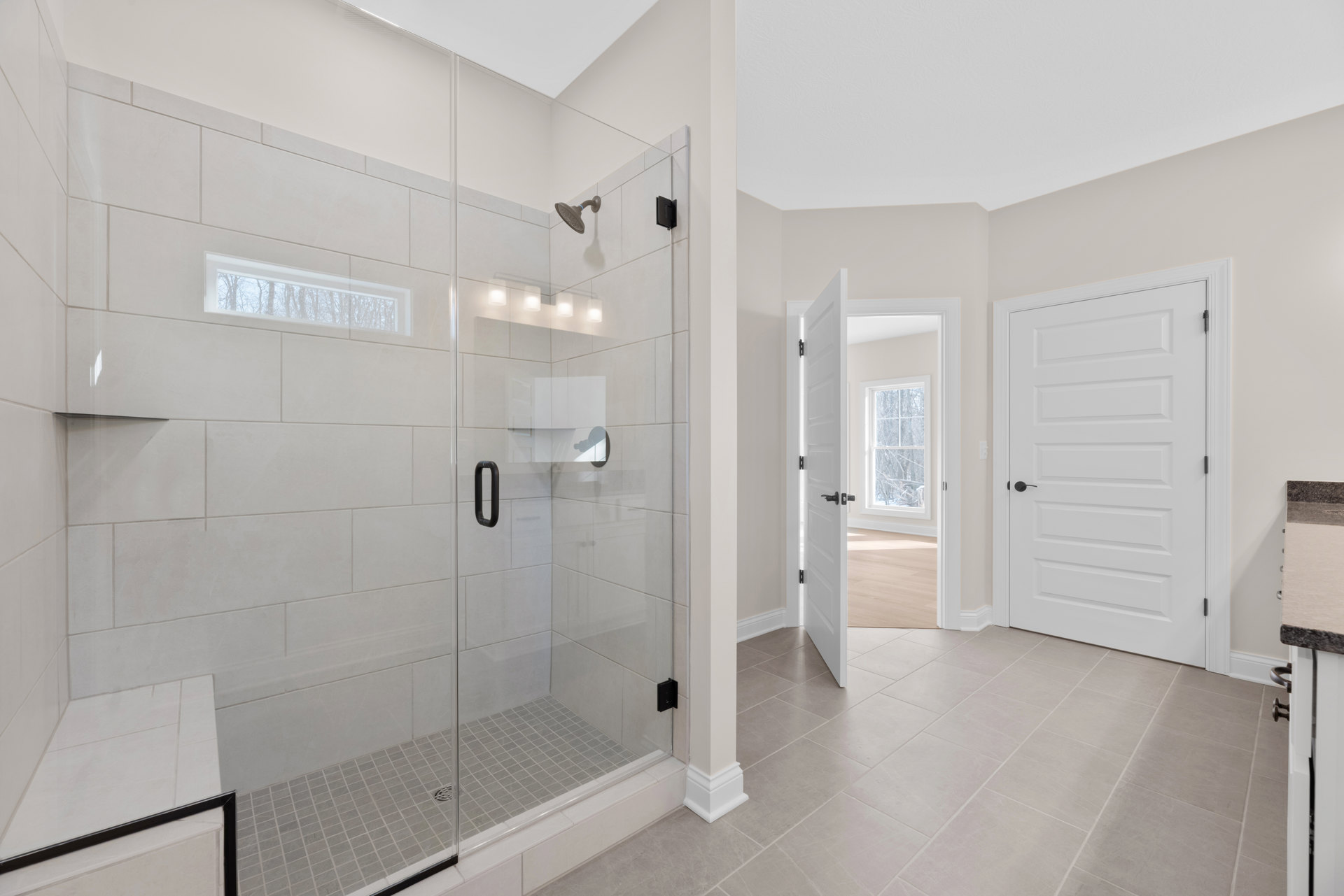 Bathroom with glass shower enclosure, white door with black handle, tile flooring, window showing trees outside, wall-mounted shower head, and modern plumbing fixtures.