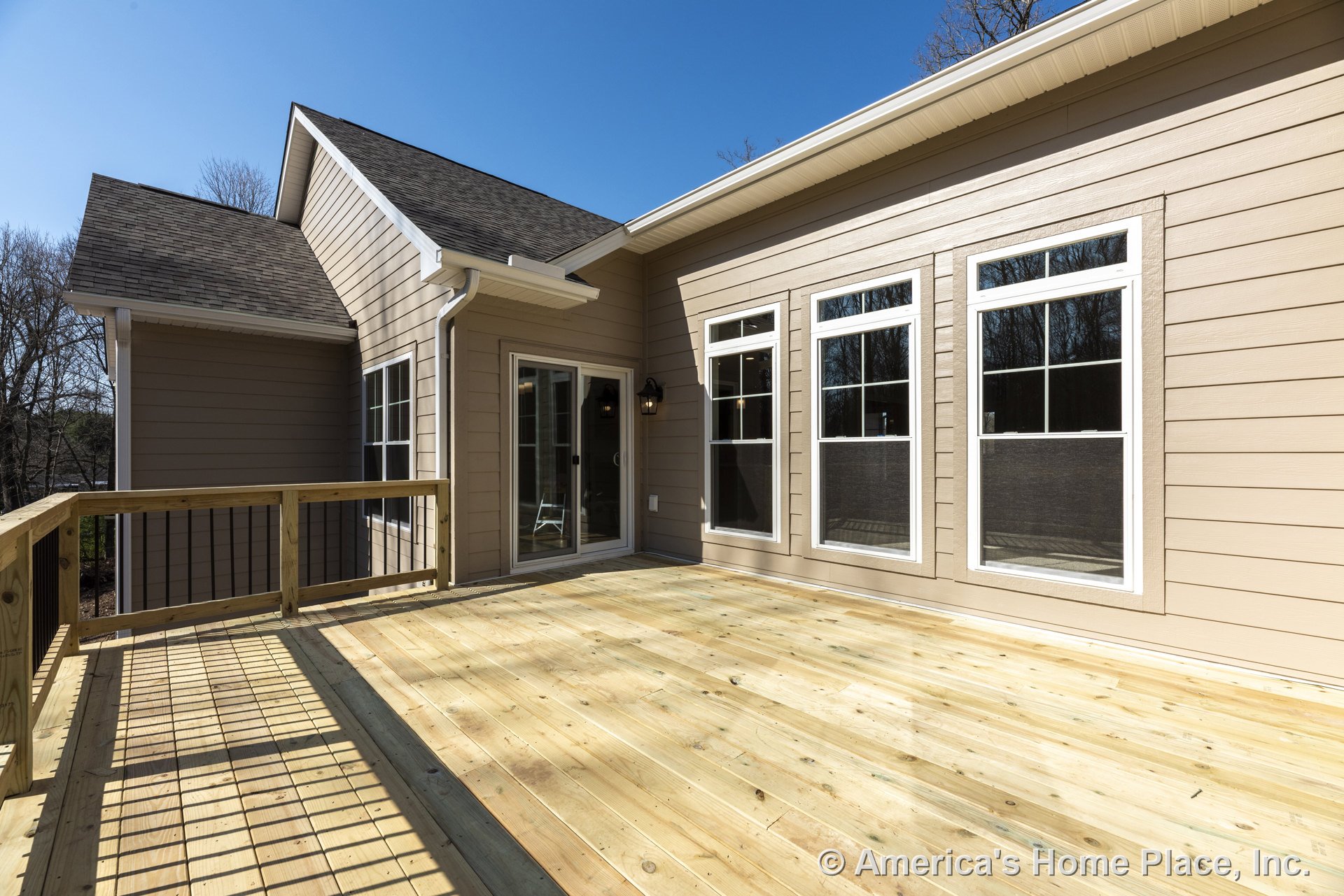 Spacious wooden deck with wood railing, beige horizontal siding, multiple large white-trimmed windows, sliding glass door, and exterior wall-mounted light fixture.