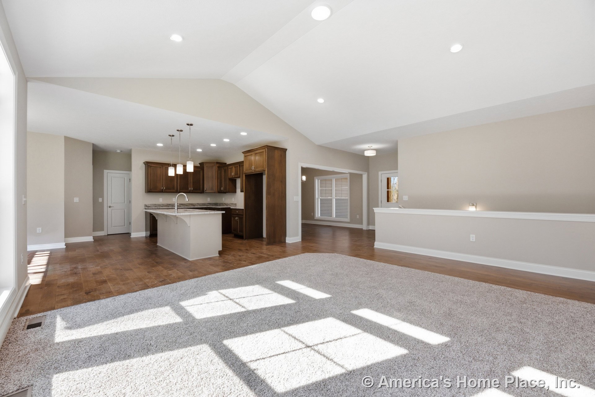 Vaulted ceiling and expansive window wall illuminate open-concept living area with wood flooring, carpeted lounge, kitchen island with sink, wood cabinetry, granite countertops