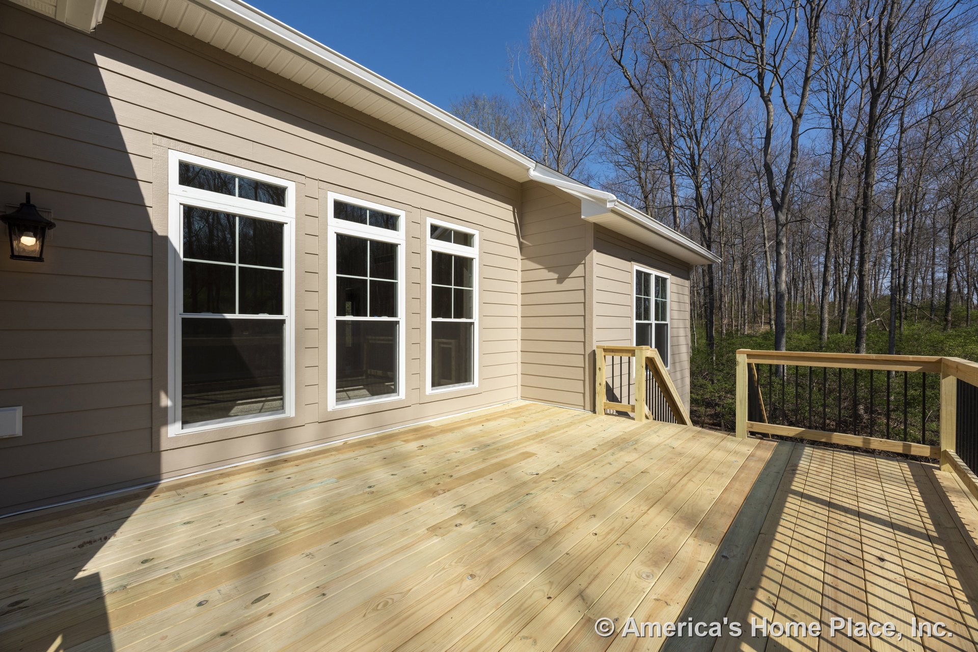 Spacious wooden deck with wood flooring and railing featuring metal balusters, attached to beige horizontal siding exterior with multiple large white-trimmed windows, black outdoor