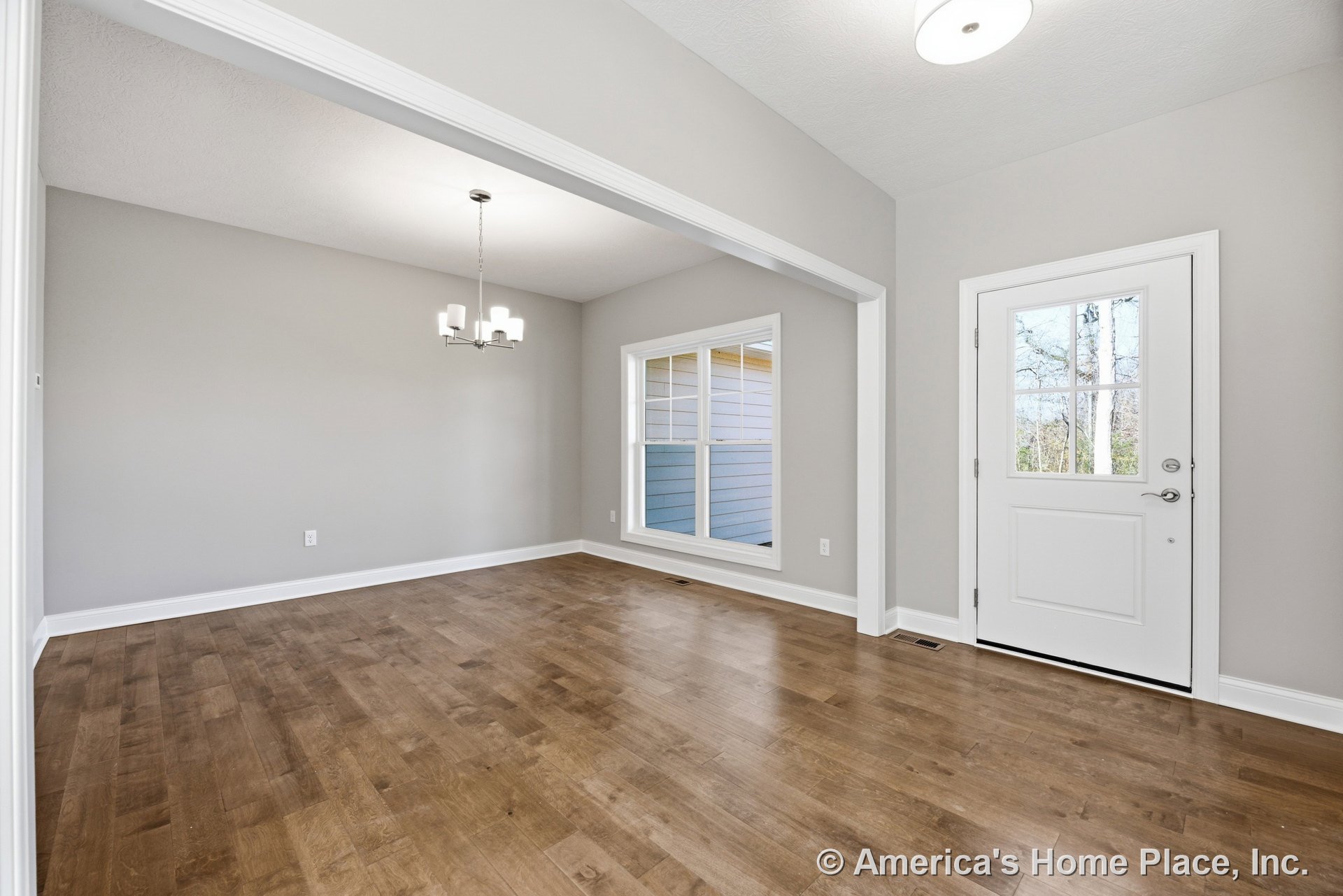 White front door with glass panes leads to open dining area featuring wood plank flooring, large double window, modern chandelier, light gray walls, and white trim with baseboards.