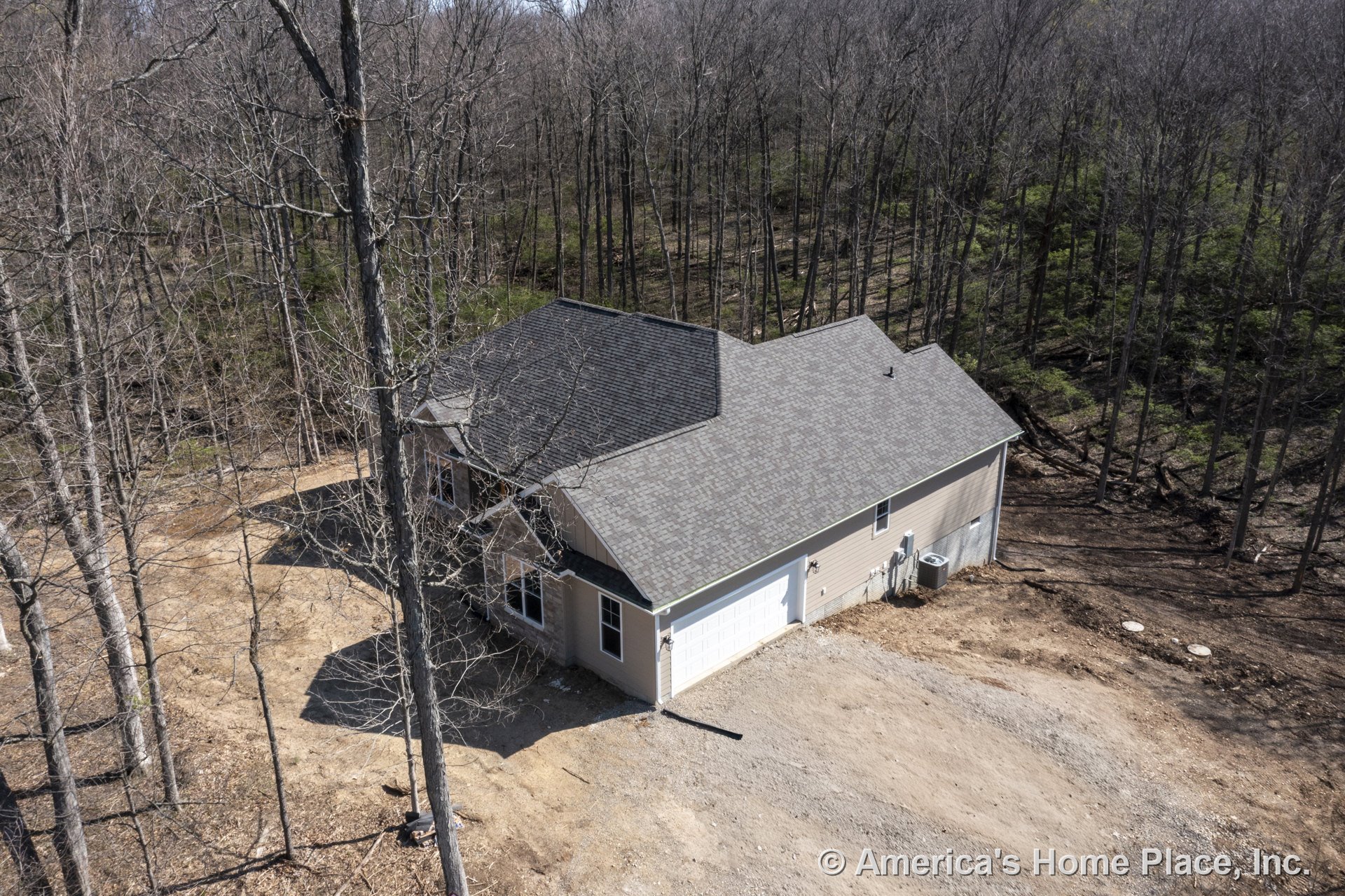 Two-car attached garage with light-colored siding, shingle roof, multiple windows, exterior lighting fixtures, and driveway set on a wooded lot; visible air conditioning unit and
