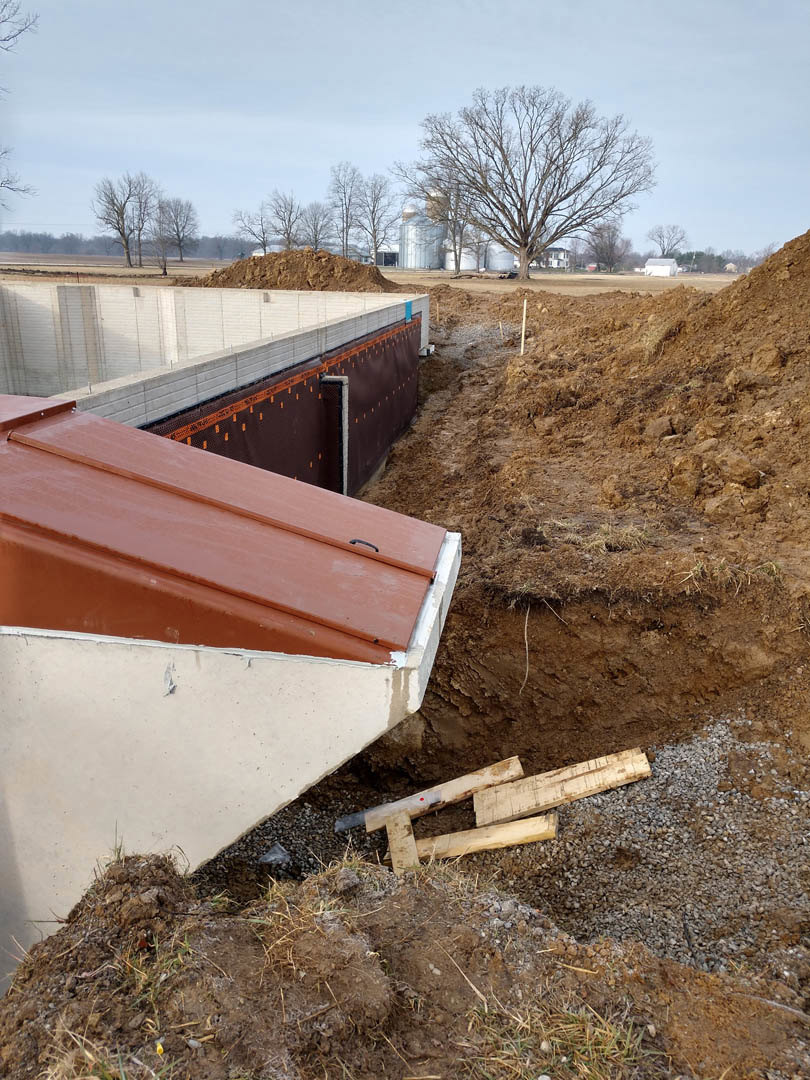 Concrete foundation slab surrounded by dirt, wooden framing materials, and leafless trees under a clear sky