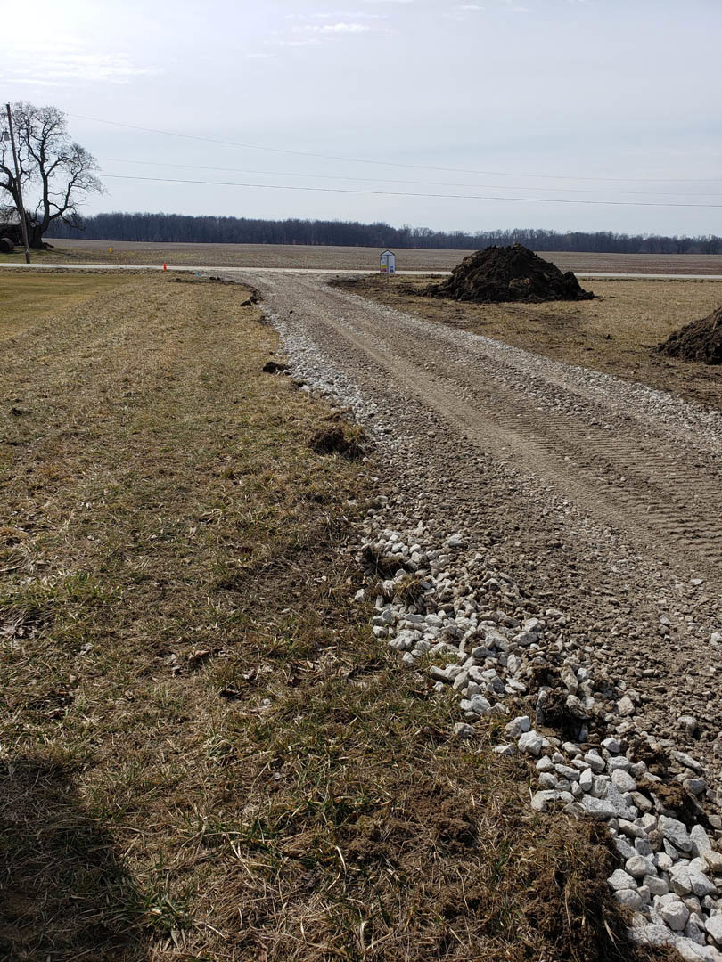 Dirt road bordered by a pile of rocks and a mound of soil, leafless tree nearby, grassy field, power lines overhead, cloudy sky