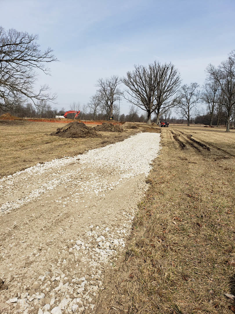 Dirt road bordered by soil piles and patches of grass, leafless trees scattered across an open field, brick wall partially visible, blue sky overhead with sparse clouds