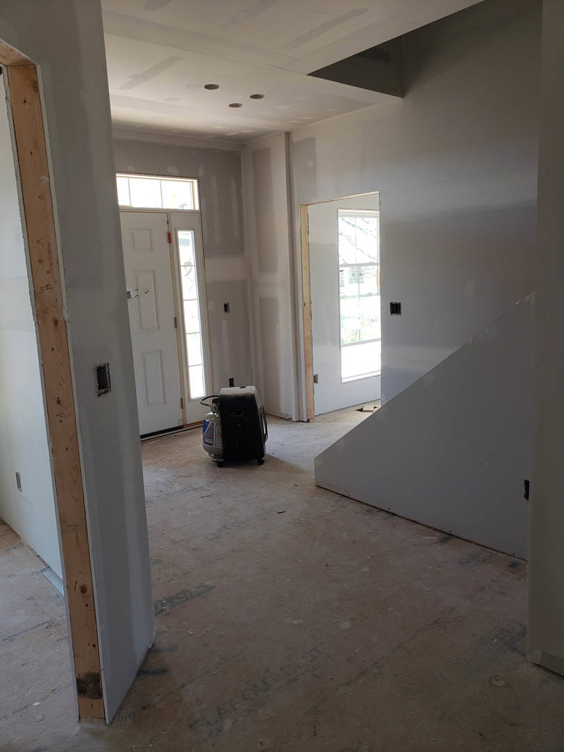 Open room with light-colored plaster walls, wood staircase with metal railing, upright vacuum cleaner near base, white door with silver knob, window overlooking street, gas