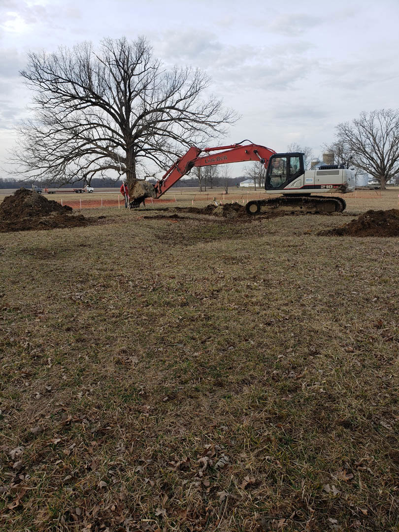 Red excavator digging into grassy field near large tree under partly cloudy sky