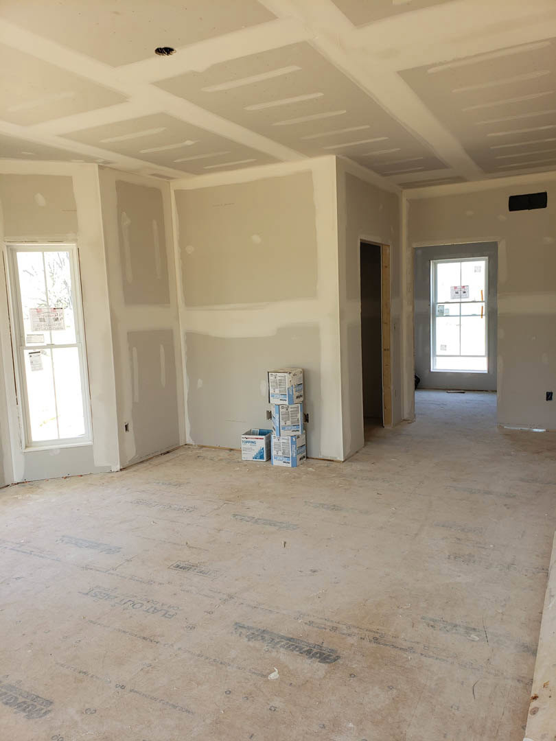 White-walled room with a window, door, and several cardboard boxes stacked on the floor and table, light-colored flooring, plaster ceiling.