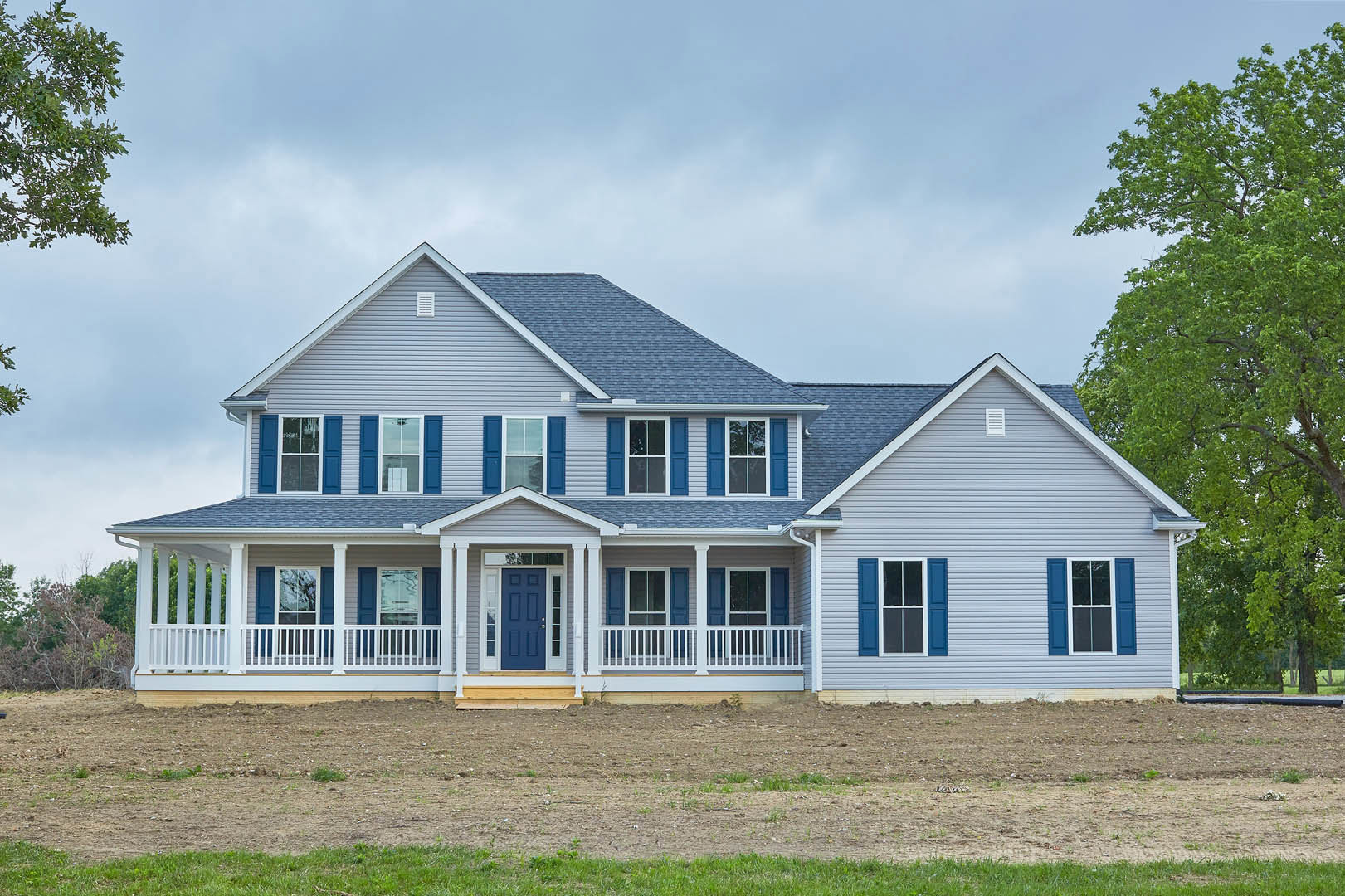 Two-story house with blue front door, white-framed windows, blue shutters, covered porch, grassy yard, and mature tree with green leaves under partly cloudy sky