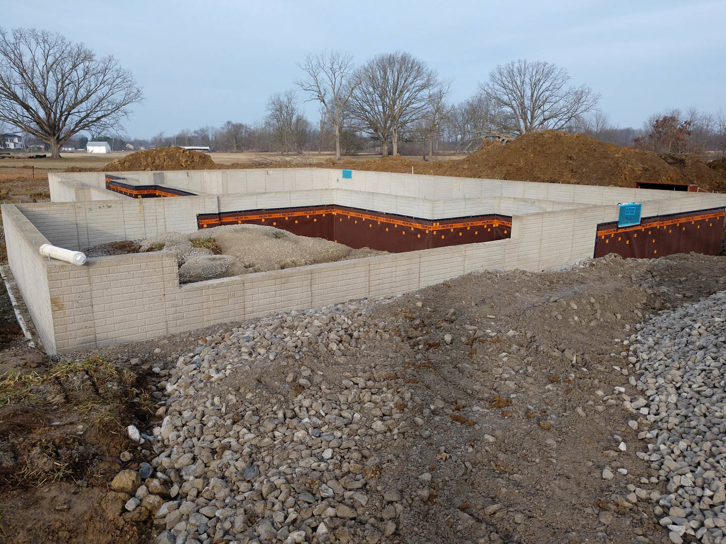 Construction site with exposed foundation, large pile of dirt and rocks, blue utility box, mature tree in grassy field, clear sky overhead