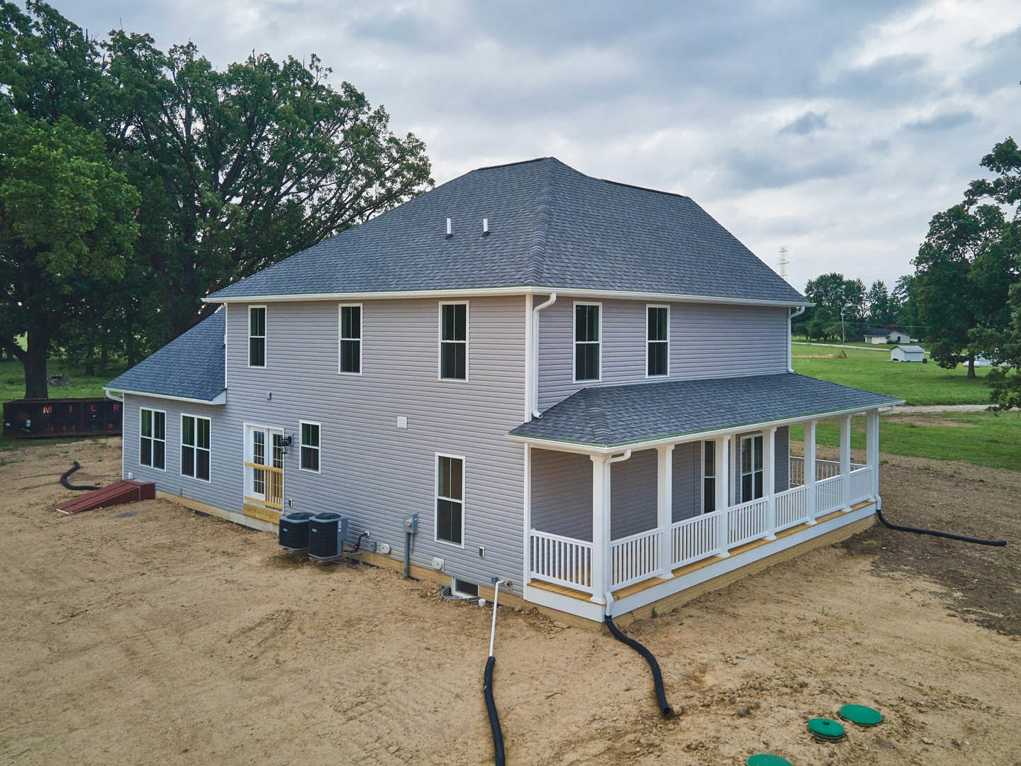 Two-story house under construction with exposed framing, large covered porch, spacious grassy yard, black and grey heat pump unit, black utility box with round top, black box with