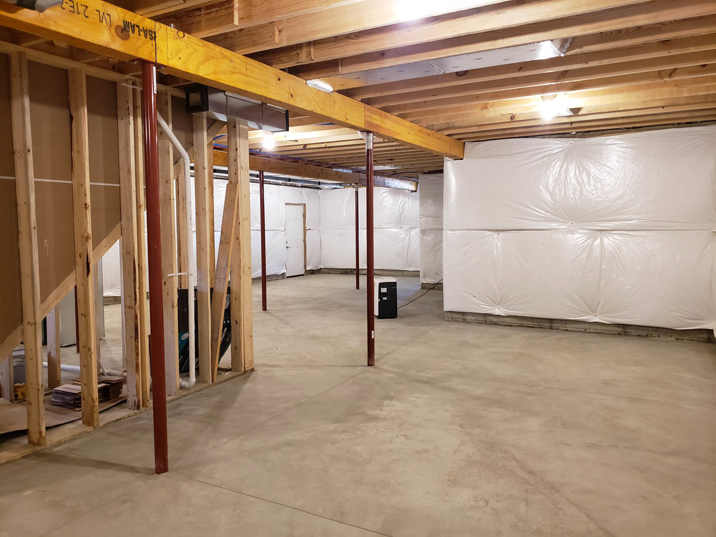 Exposed wooden ceiling beams, white plastered wall with plastic sheeting, unfinished concrete floor with red support poles, black square frame mounted on wall, basement