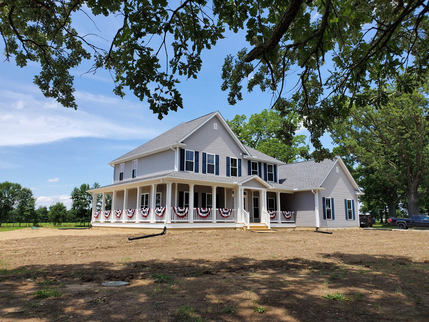 Two-story home with covered front porch, white railing, large tree in yard, dirt field, and cloudy sky in background