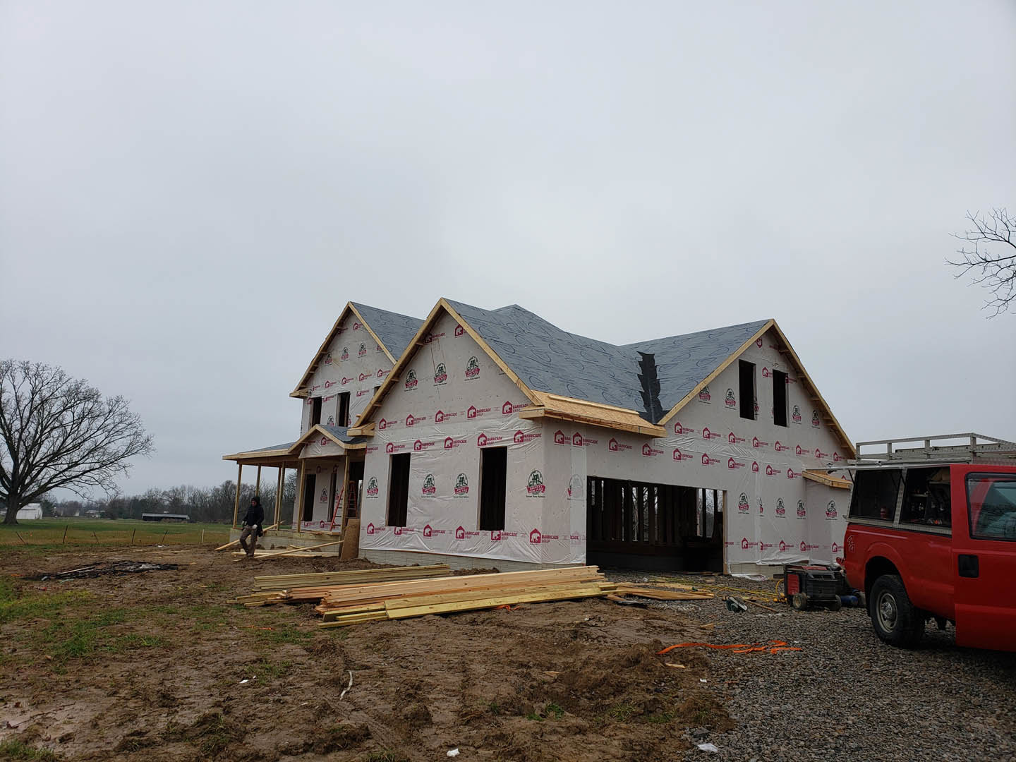 Partially built house with plastic sheeting, leafless tree, red truck with ladder, wood planks scattered on ground, overcast sky