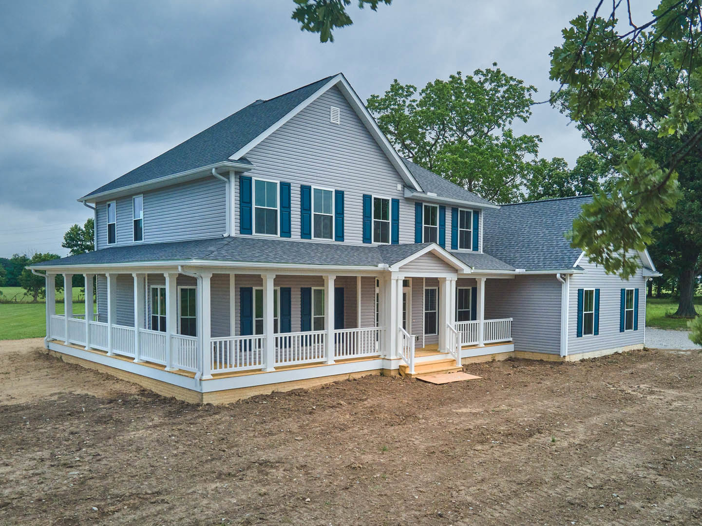 Large front porch with white railings, multiple windows with white frames, dirt patch in front, surrounded by trees under a partly cloudy sky.