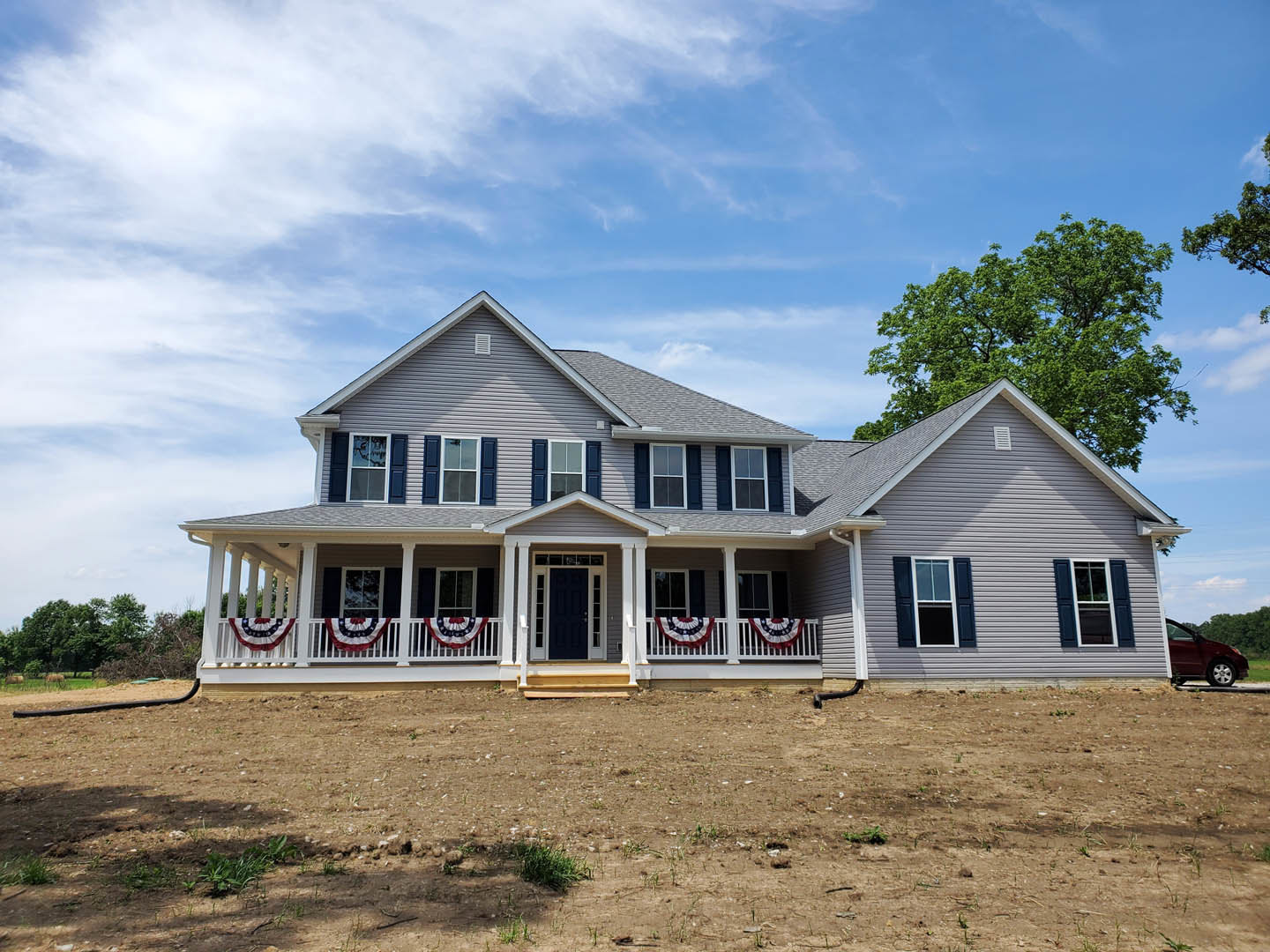 Two-story house with white trim and black front door, covered porch with white railing, large tree in front yard, cloudy sky overhead, car parked beside yard sign.