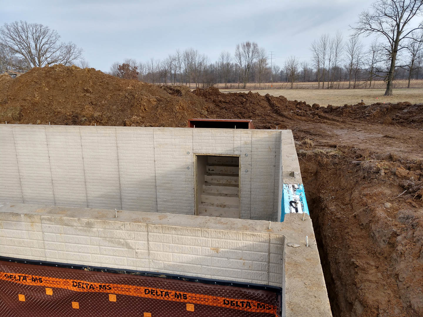 Concrete foundation with exposed hole in the ground, surrounded by bare earth and a row of trees under a clear sky