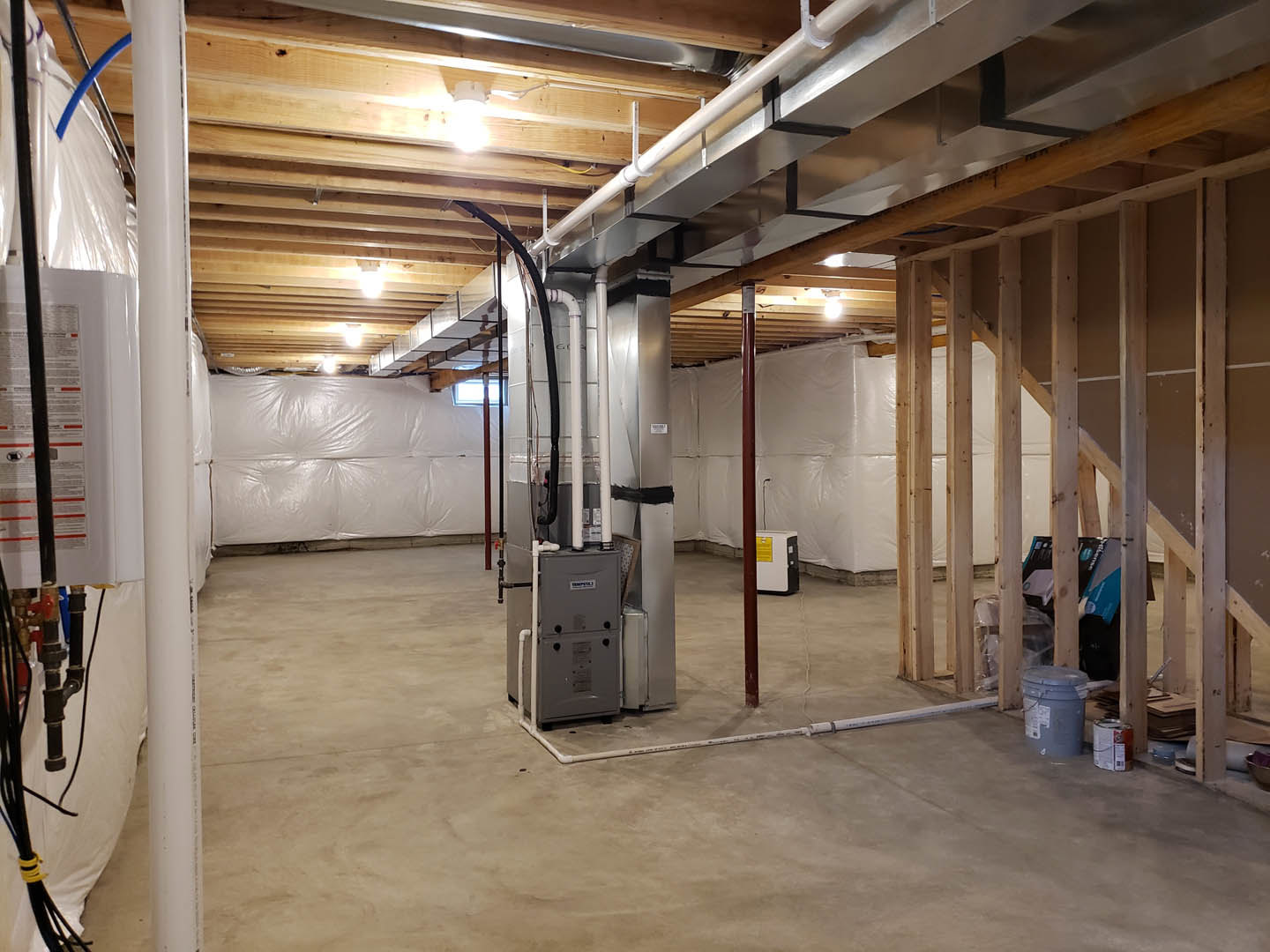 Unfinished basement with exposed steel beams, concrete floor, large metal utility box, visible pipes, white plastic wall covering, grey bucket with lid, and bright wall-mounted