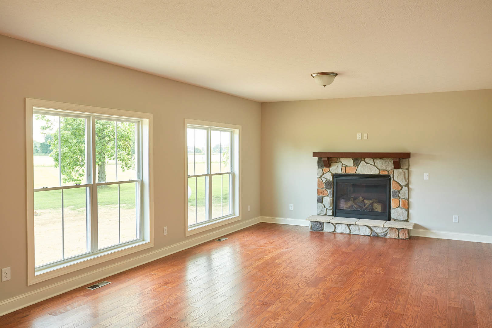 Living room with hardwood floors, stone fireplace with wood logs burning, large windows showing leafy tree outside