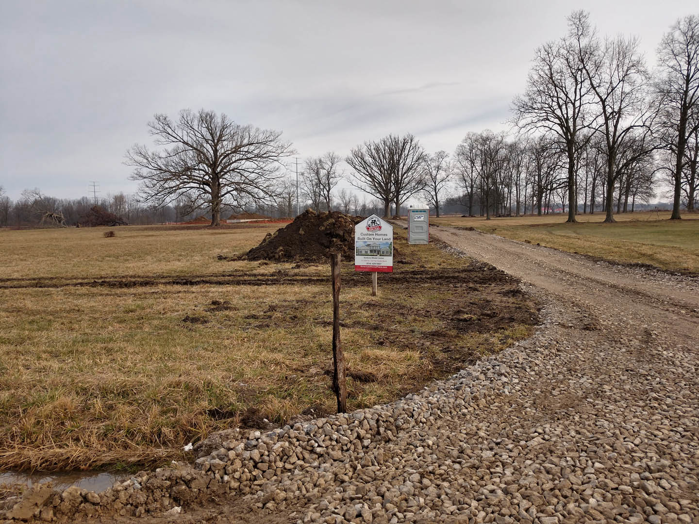 Dirt road leading through grassy field with a sign displaying a house illustration, large leafless tree and group of bare trees in background, pile of dirt beside sign under cloudy