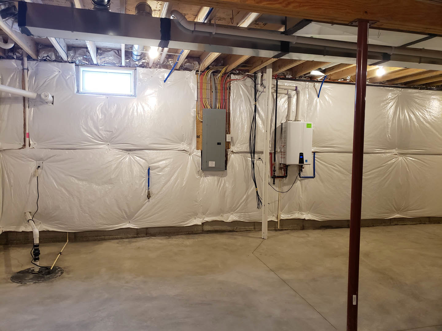 Basement room with concrete floor, white plastic covering draped over surfaces, grey door with square black handle, exposed beams, white refrigerator with green label, and visible