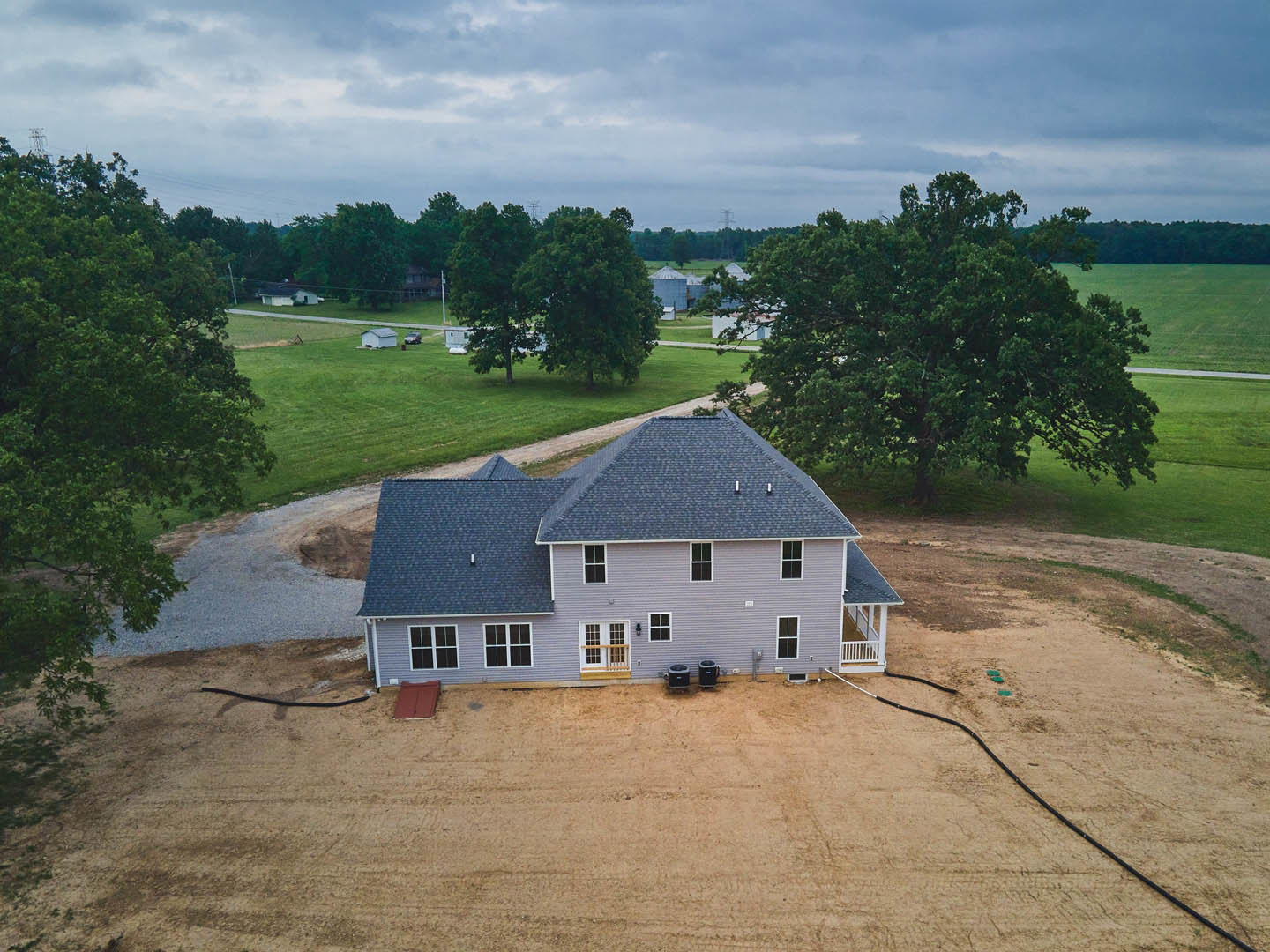 Two-story house with blue roof, white siding, large paved driveway, mature trees, grassy lawn, and cloudy sky