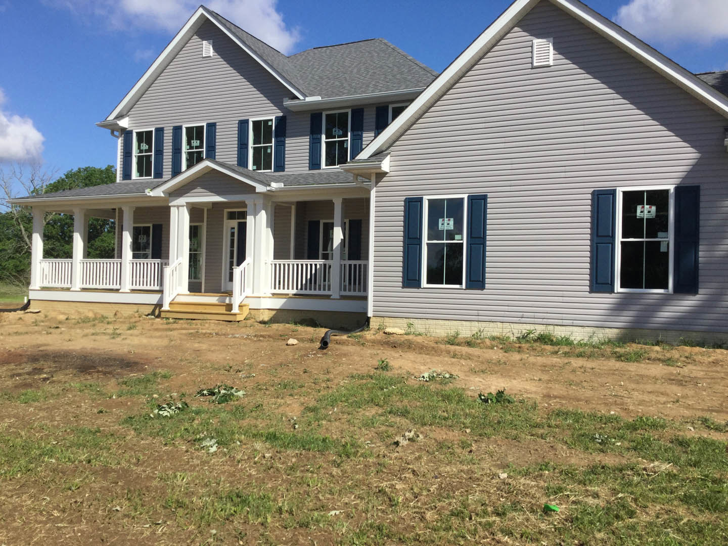 Two-story house with covered porch and upper balcony, black pipe extending from exterior wall, grassy yard bordered by dirt field, window displaying a sign, white clouds in blue