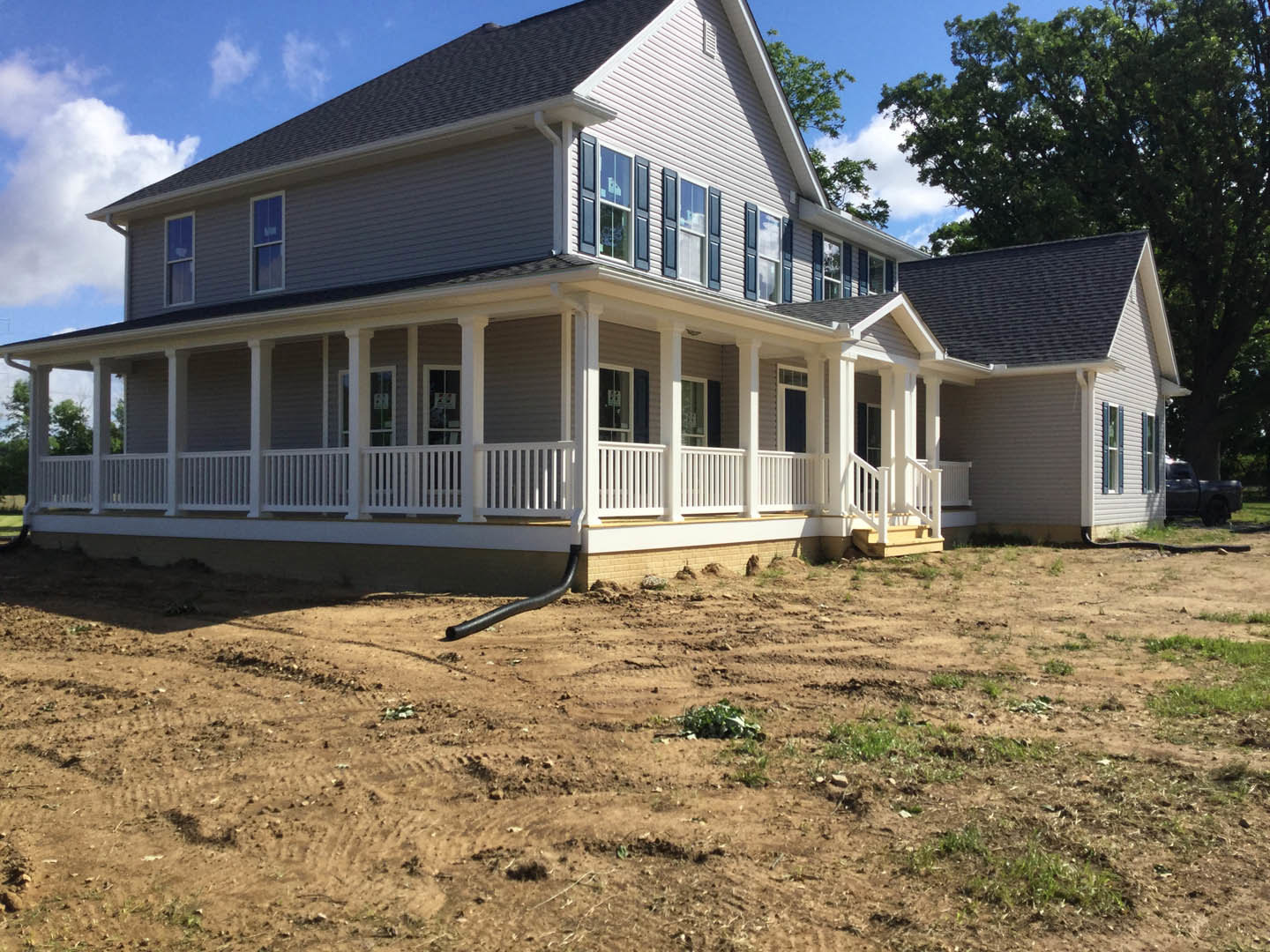 Large front porch with white railing, light-colored siding, multiple windows, tree in background, dirt field with black pipe, parked truck near utility pole