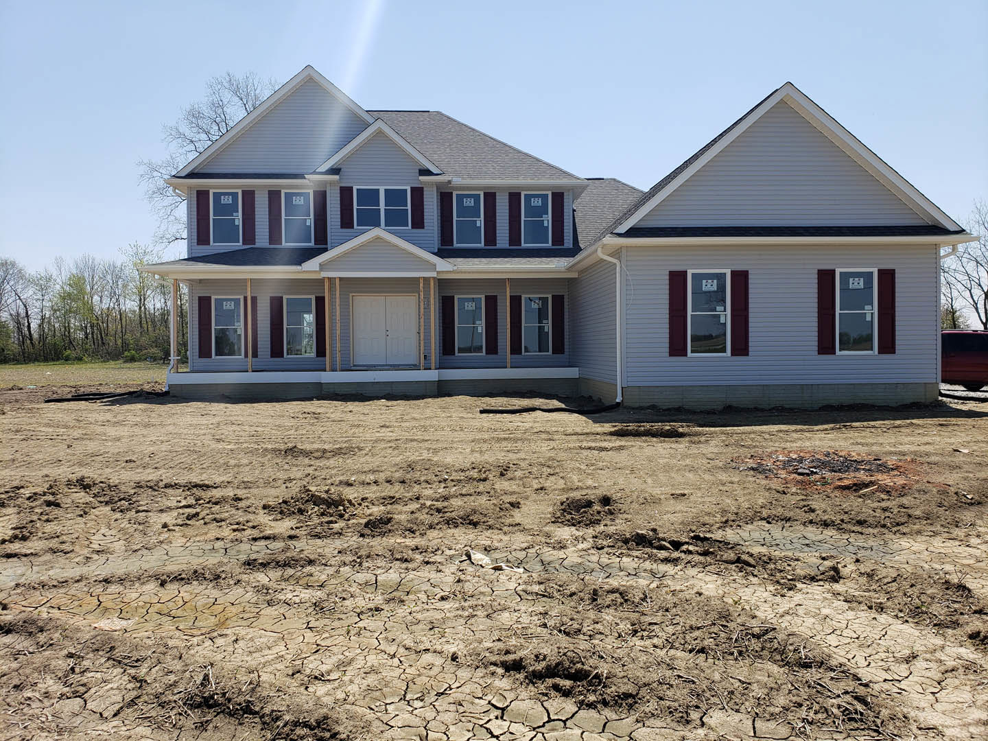 Modern house under construction with exposed dirt yard, double entry doors, light-colored siding, large windows, and a red car parked along the street under a clear blue sky