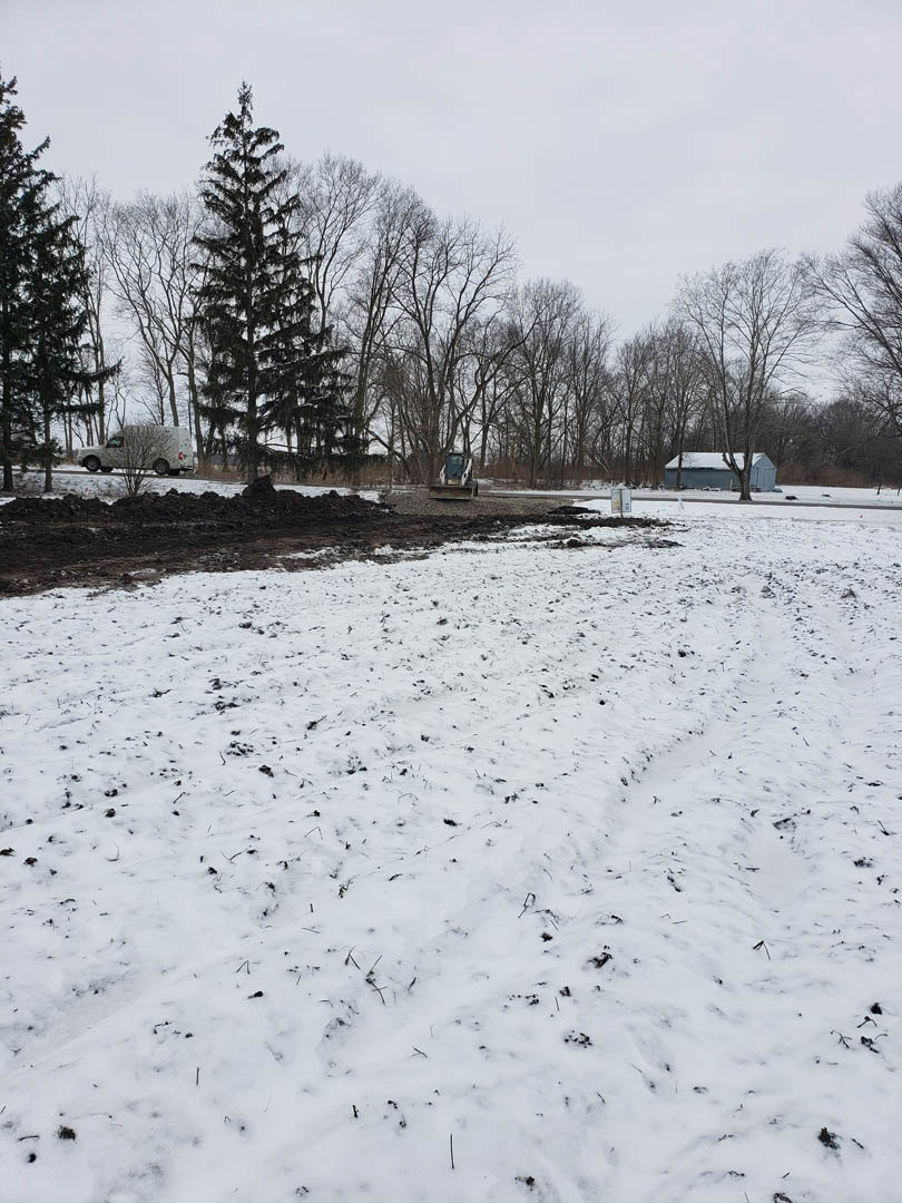 Snow-covered field with scattered trees, a tractor parked near a white van, and a building partially visible in the background