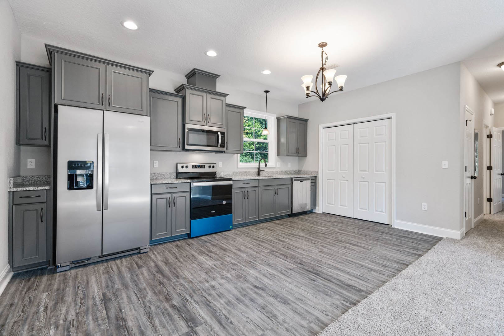 Grey kitchen cabinets with brushed metal hardware, wood plank flooring, stainless steel refrigerator and stove, white interior door, modern chandelier above island, black and blue