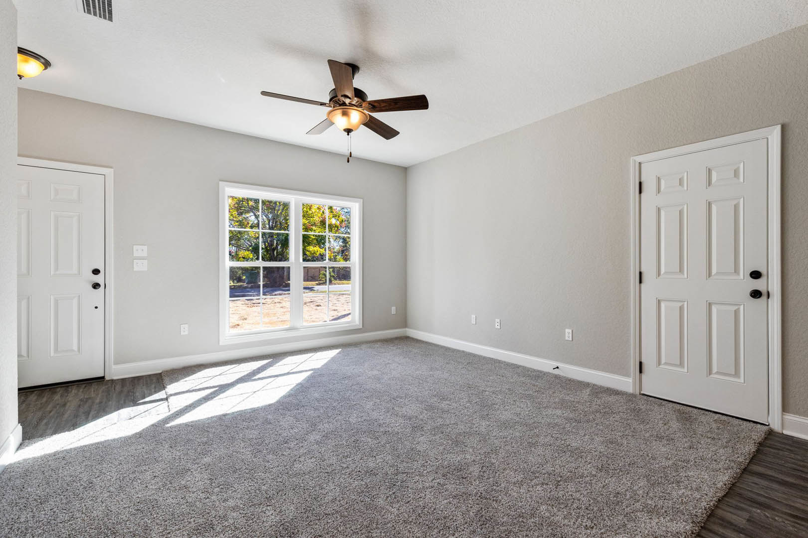 Bedroom with carpet flooring, ceiling fan with light fixture, large window overlooking trees, white paneled door with black hardware, neutral painted walls