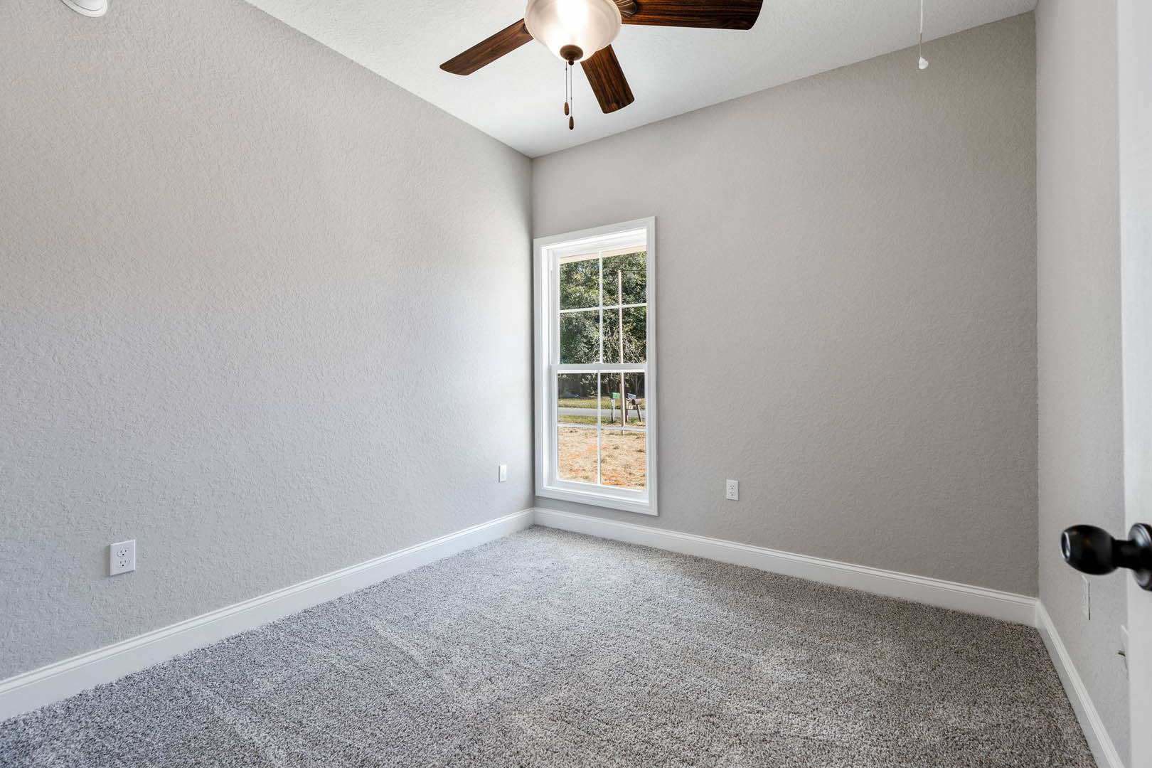 Neutral-toned room with carpet flooring, white walls, ceiling fan with light fixture, large window overlooking green yard, white door with brushed metal knob, simple baseboard