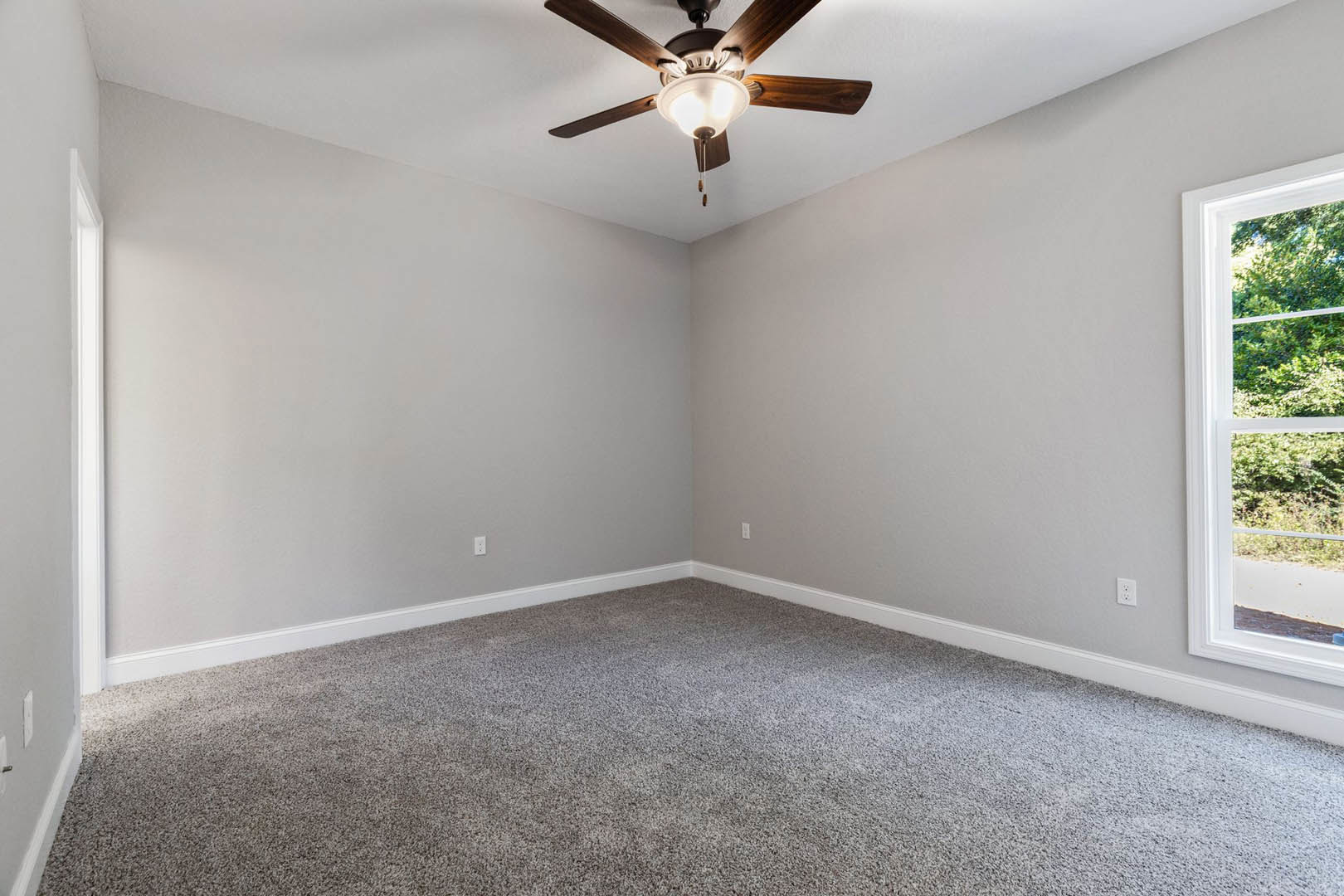 Neutral-toned carpeted bedroom with white walls, ceiling fan featuring light fixture, large window overlooking leafy trees, simple crown molding along ceiling edges