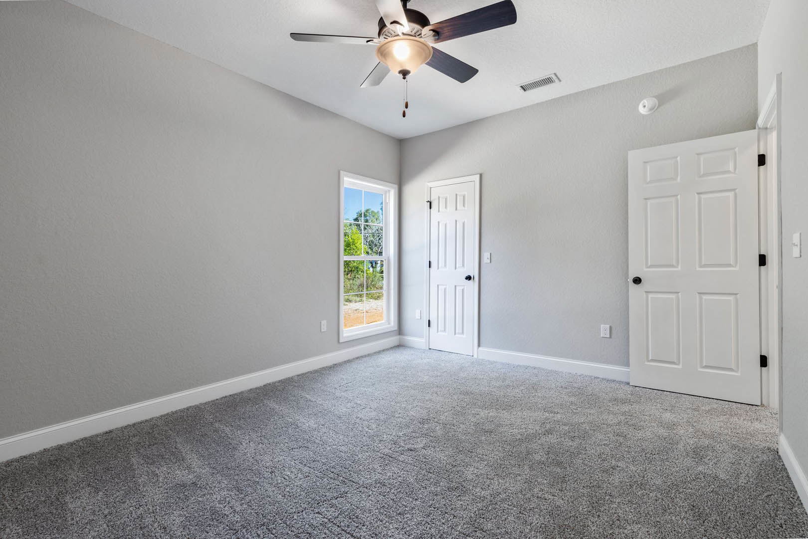 Carpeted room with white walls, ceiling fan with light fixture, large window showing trees, and white door with black hardware