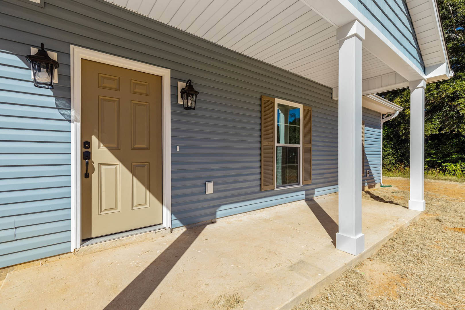 Blue siding exterior with white-trimmed porch, paneled front door, shuttered window, glass wall sconce, and concrete patio