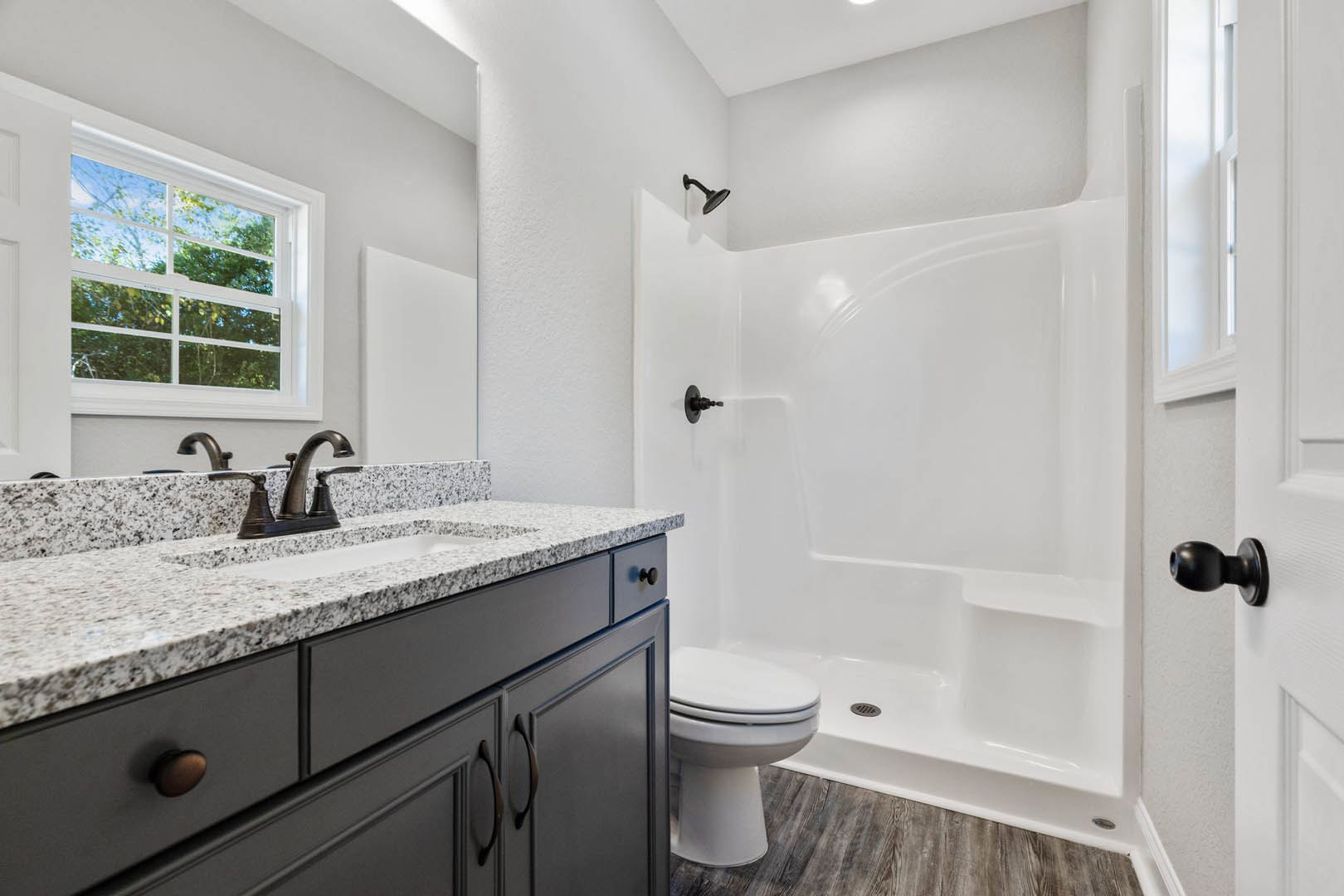 Bathroom with white tile shower, glass enclosure, white porcelain sink, stone countertop, modern faucet, closed toilet, window overlooking trees, light cabinetry, and chrome