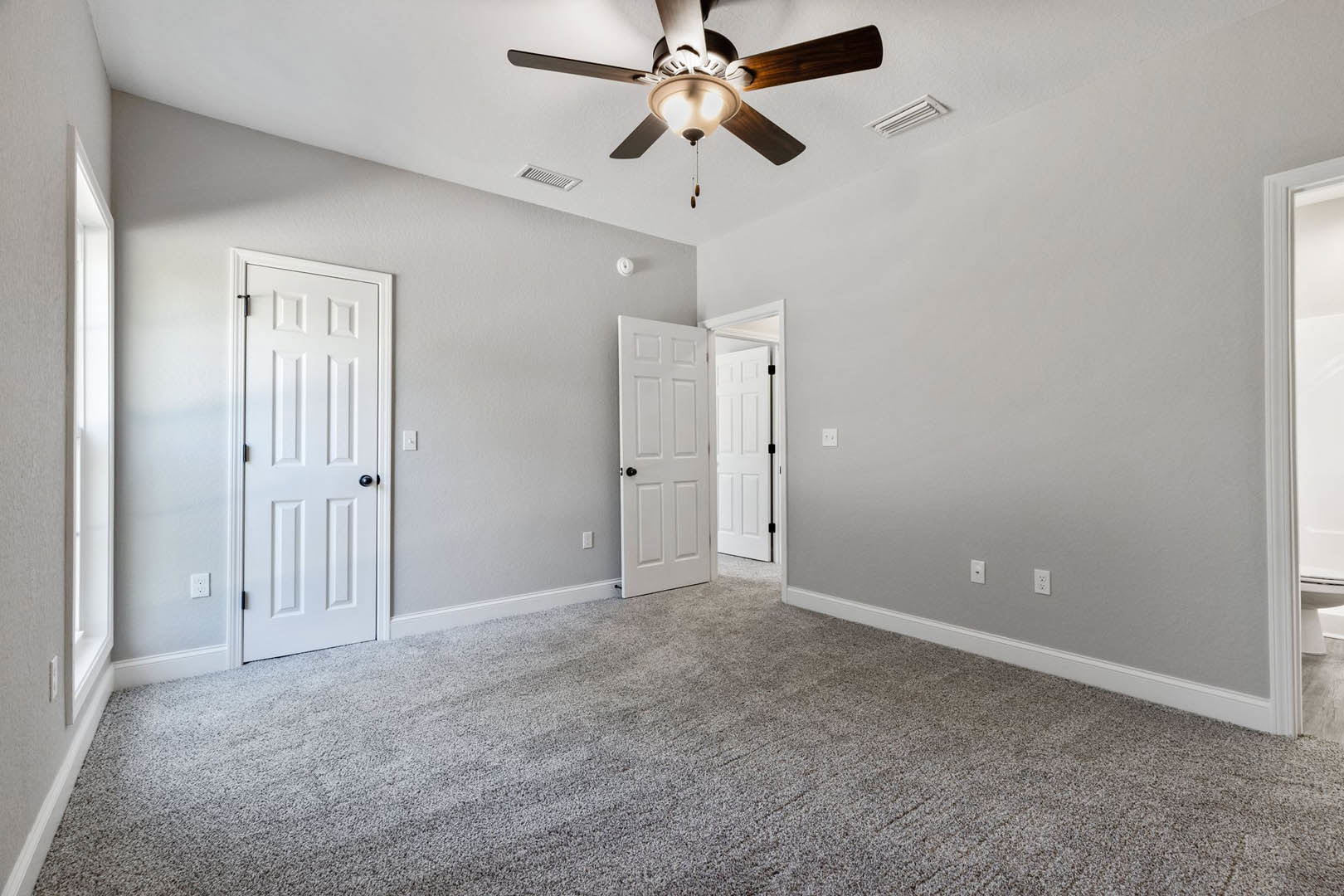 Carpeted room with white walls, ceiling fan with light fixture, two white doors featuring black hardware, and a white vent cover on the wall