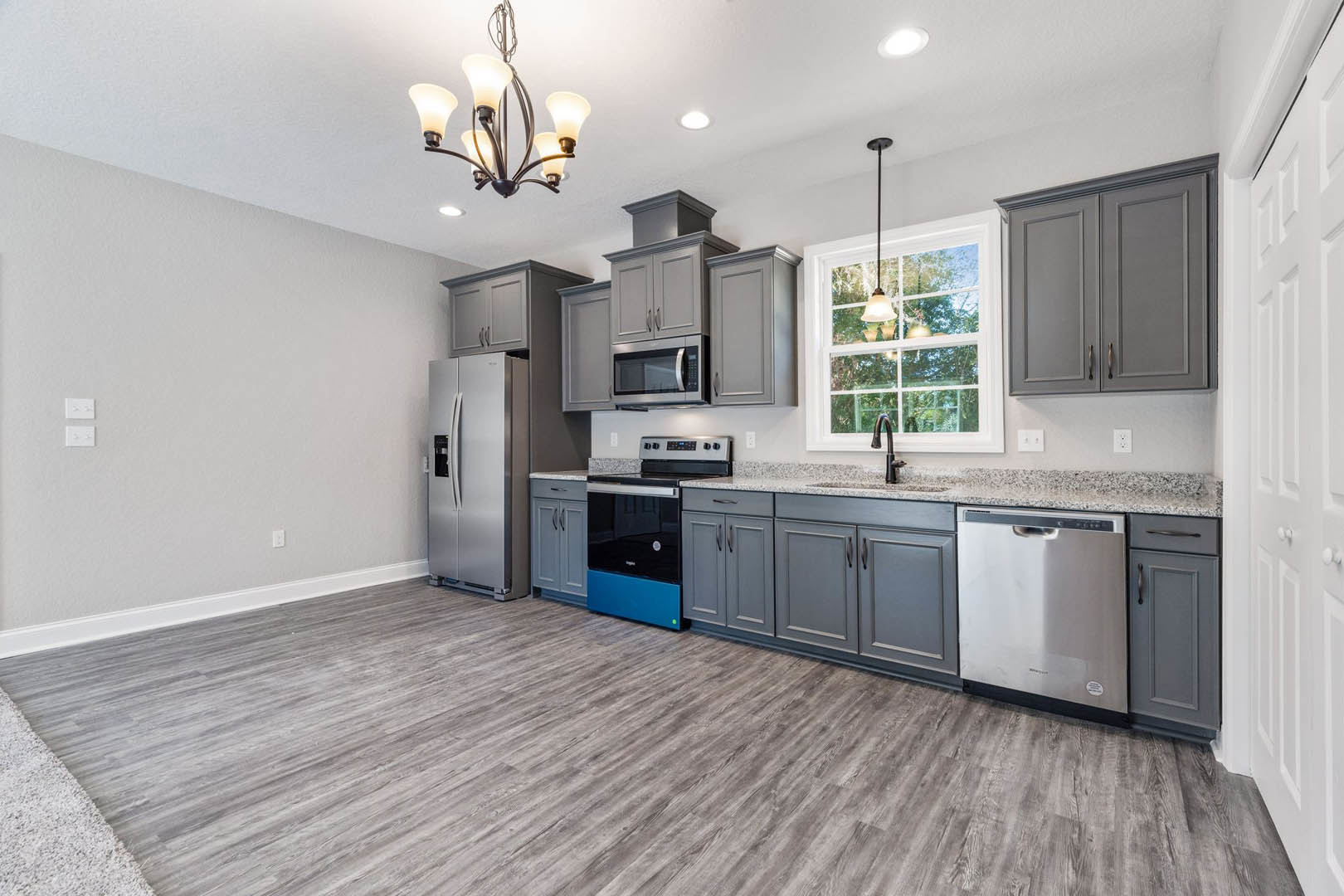 Grey kitchen cabinets with brushed metal handles, wood plank flooring, stainless steel refrigerator and dishwasher, five-light chandelier, black and blue oven and drawer, white