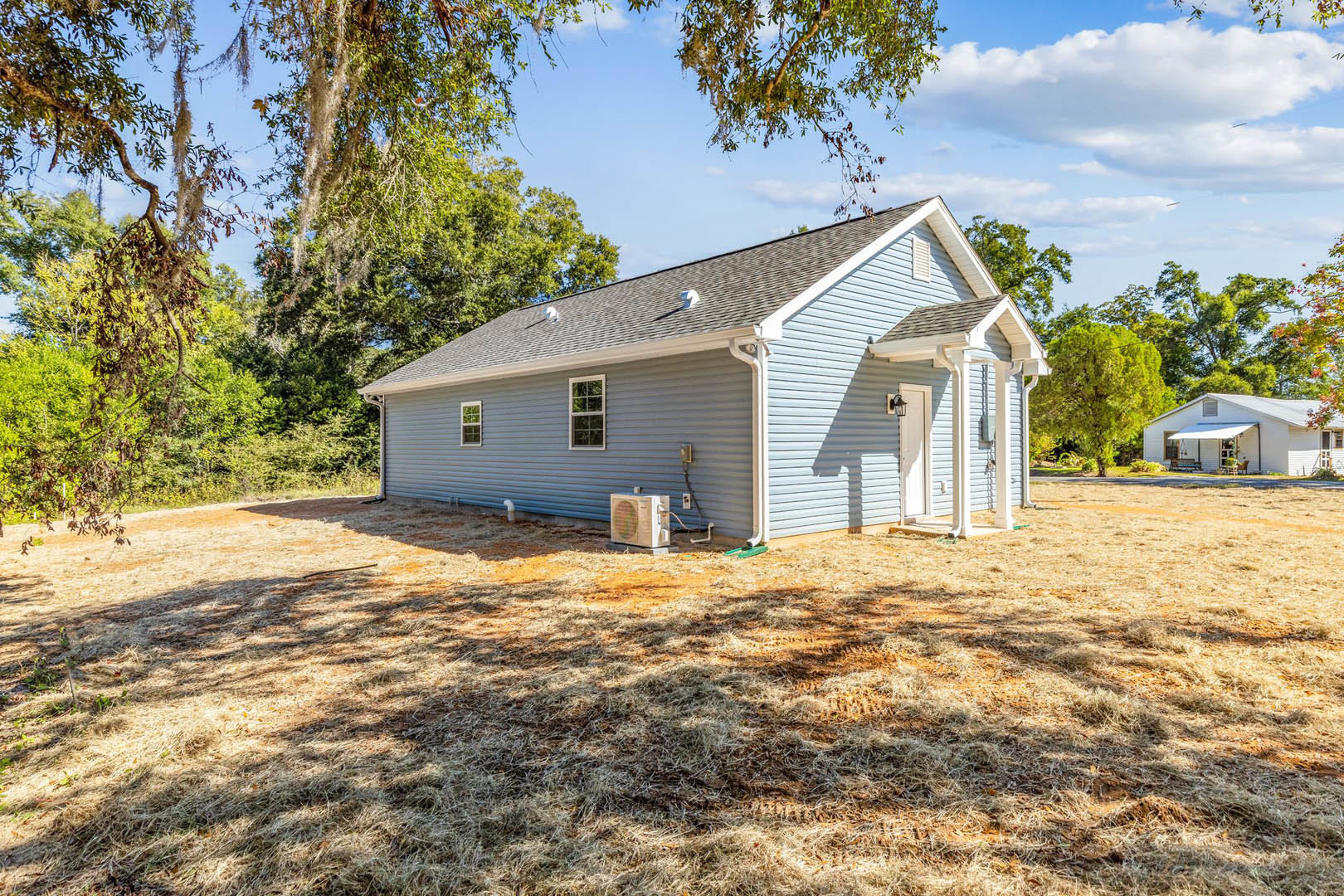 White single-story house with white roof, white-framed windows, white awning, fenced yard, outdoor heater, grassy lawn, and mature trees in the background