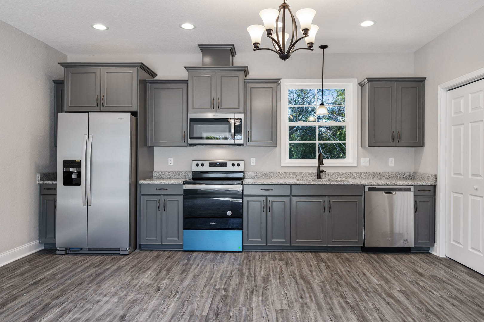 Grey kitchen cabinets with brushed metal hardware, wood plank flooring, stainless refrigerator, black and blue oven, built-in microwave, quartz countertops, window with natural