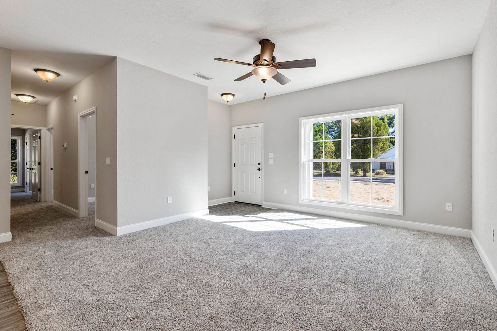 Carpeted bedroom with white walls, ceiling fan with light, large window overlooking street and trees, white door with black hardware