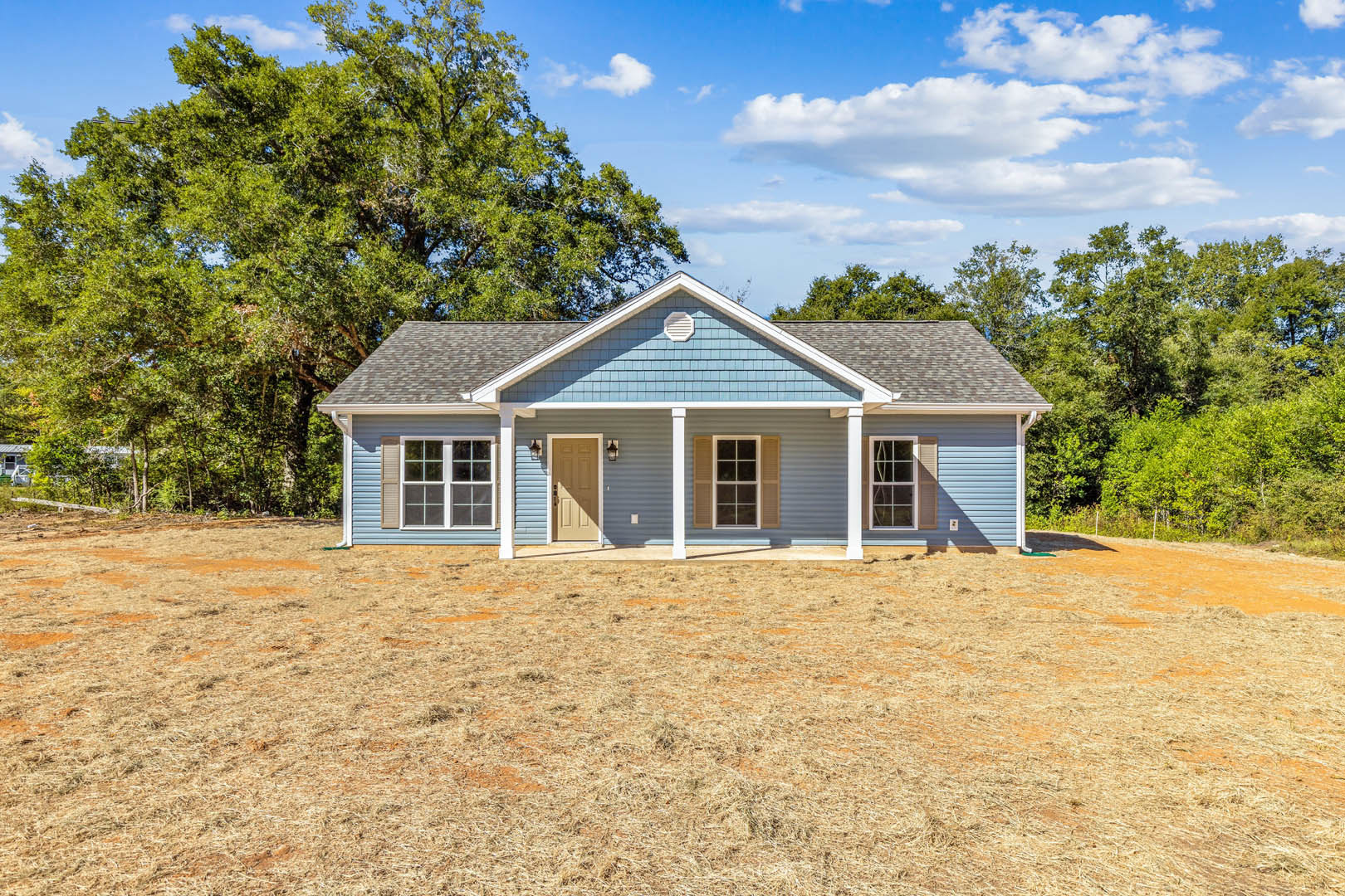 Blue siding house with white columns, white front door with door light, shuttered windows, large grassy yard, mature tree with green leaves, clear blue sky