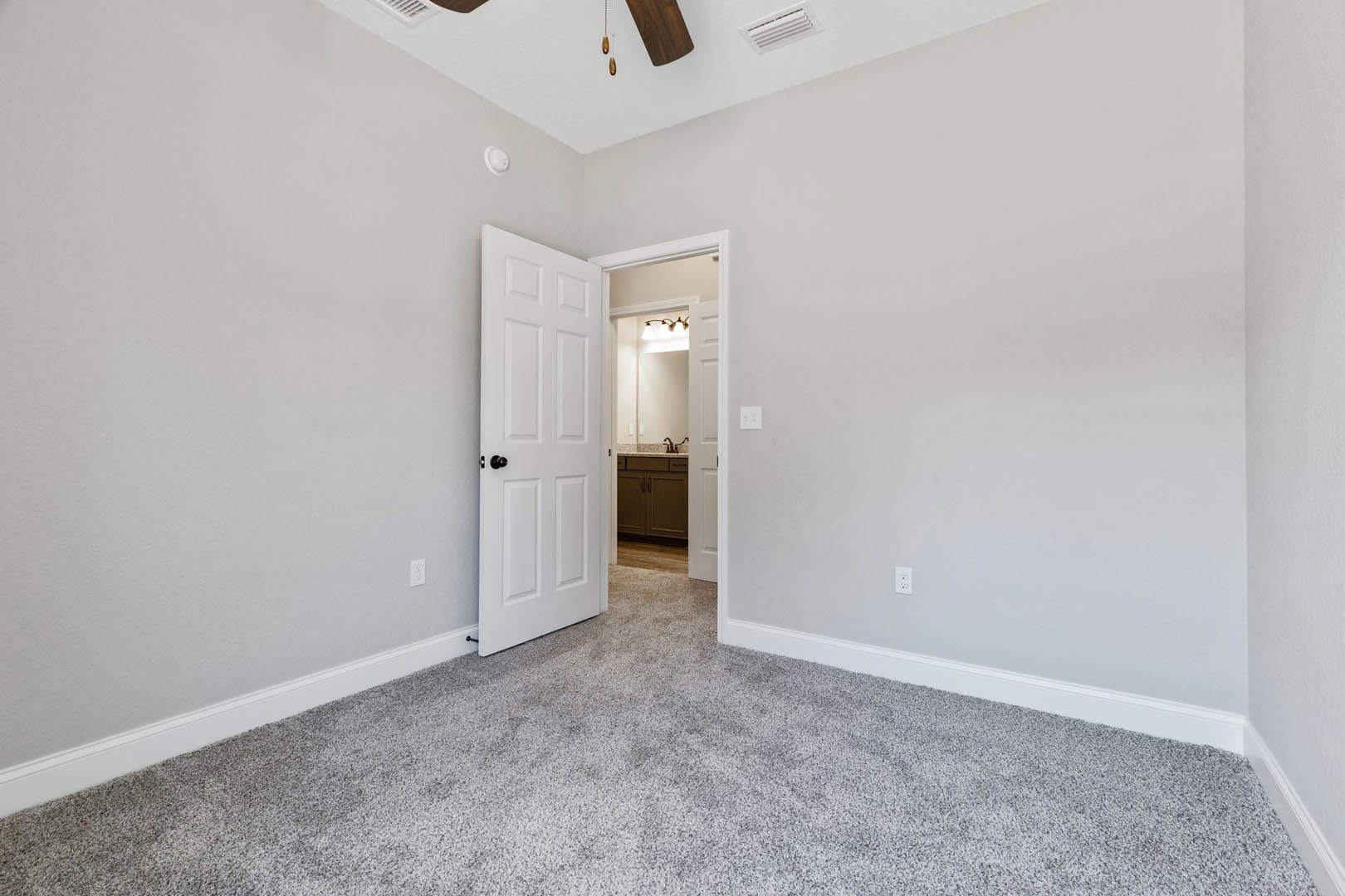 Carpeted bedroom with white baseboards, ceiling fan, white door with black knob, adjacent bathroom featuring a mirror and wood cabinet