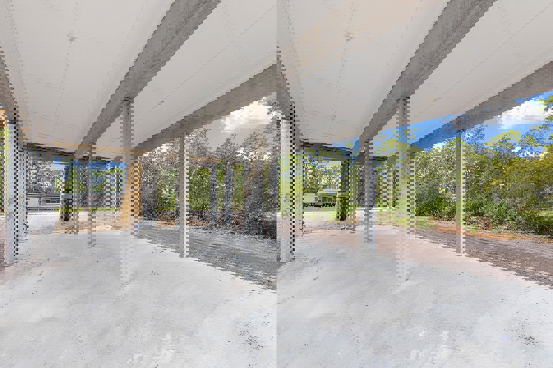 Concrete structure with exposed pillars, unfinished exterior, dirt road in foreground, surrounding trees, partially constructed ceiling beams